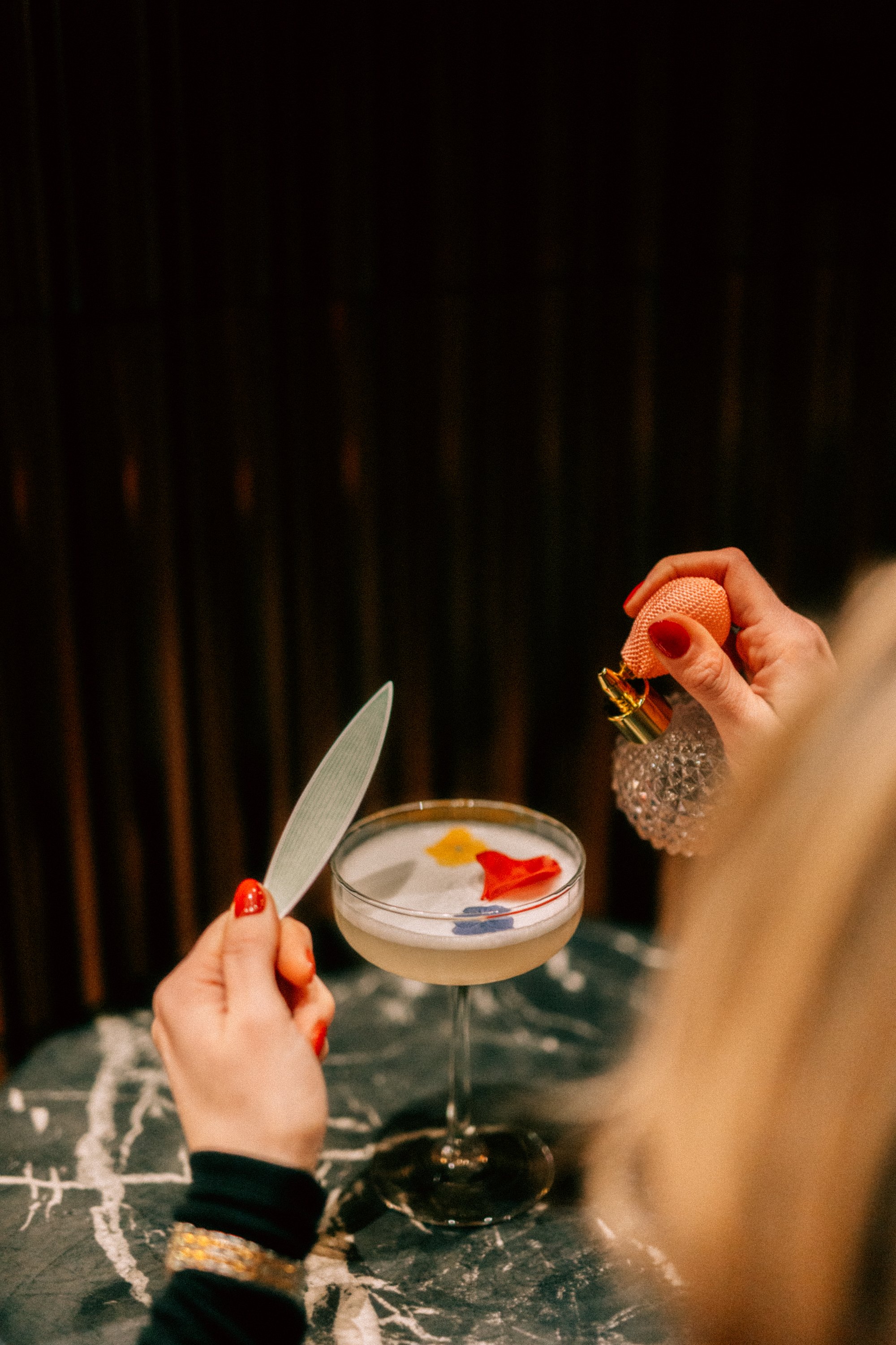 Close-up of a person holding a perfume atomizer in one hand and a knife in the other, while preparing to spray perfume onto a cocktail glass filled with a drink garnished with colorful flower petals. The person has red nail polish and is wearing a black long-sleeve shirt with a gold bracelet. The background is dark and the surface is a black marble table.