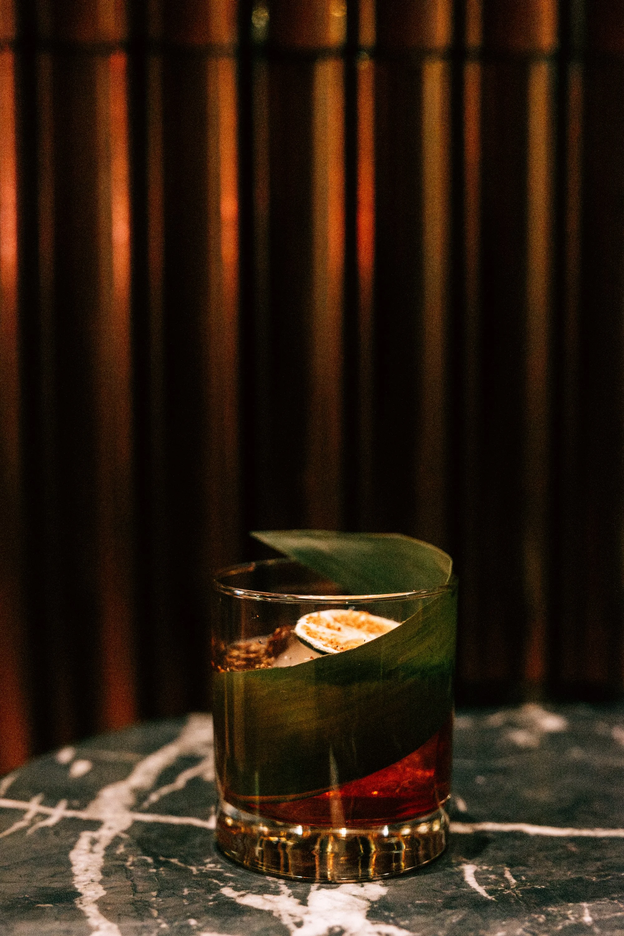 A glass of dark-colored cocktail with a lime wedge and ice, placed on a black marble surface with white veining, against a dark background with vertical lines.