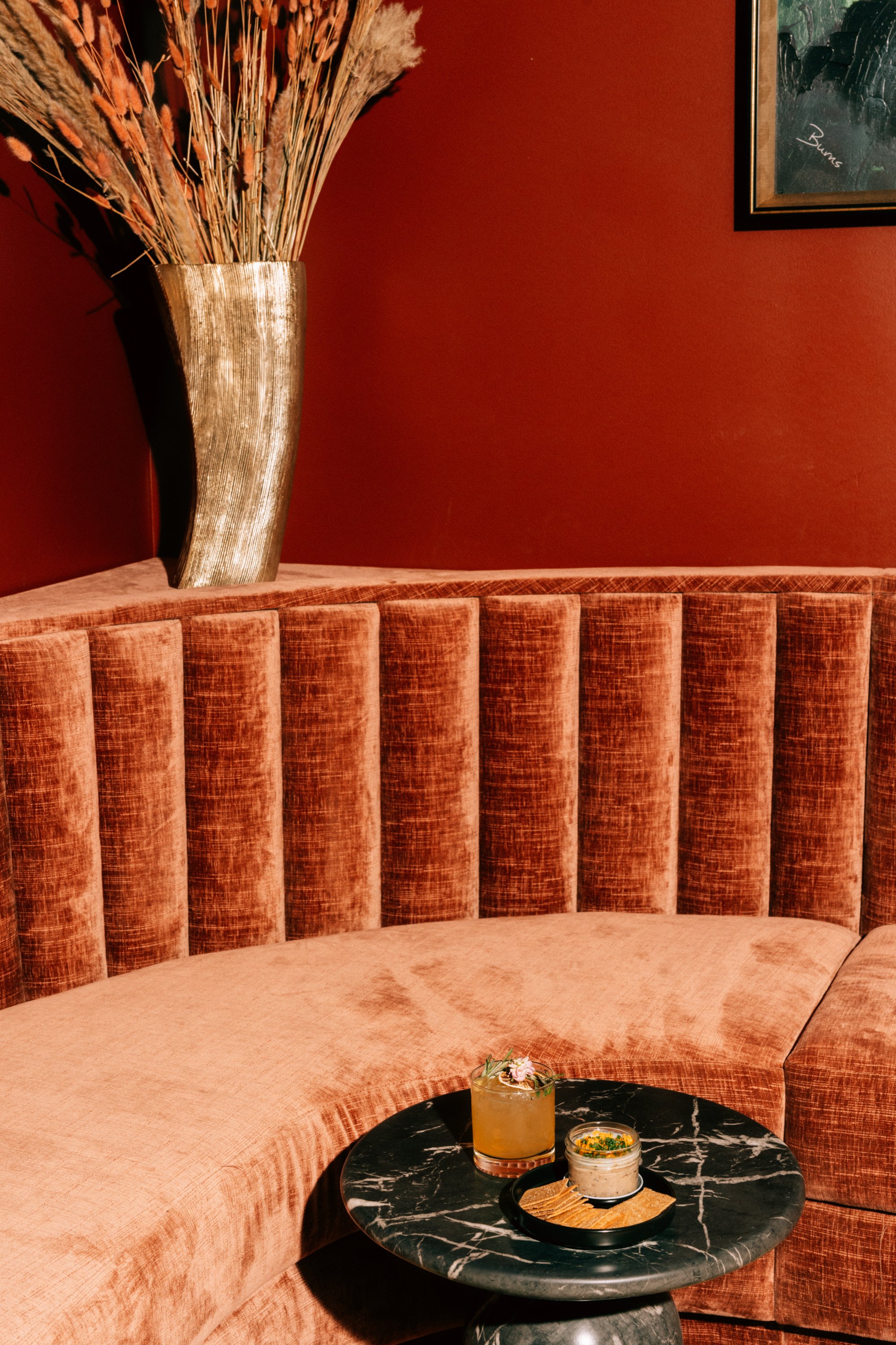 A corner of a room with a plush, pink velvet curved sofa, a black marble side table with drinks, a tall gold vase with dried flowers, and a dark red wall with a framed picture.