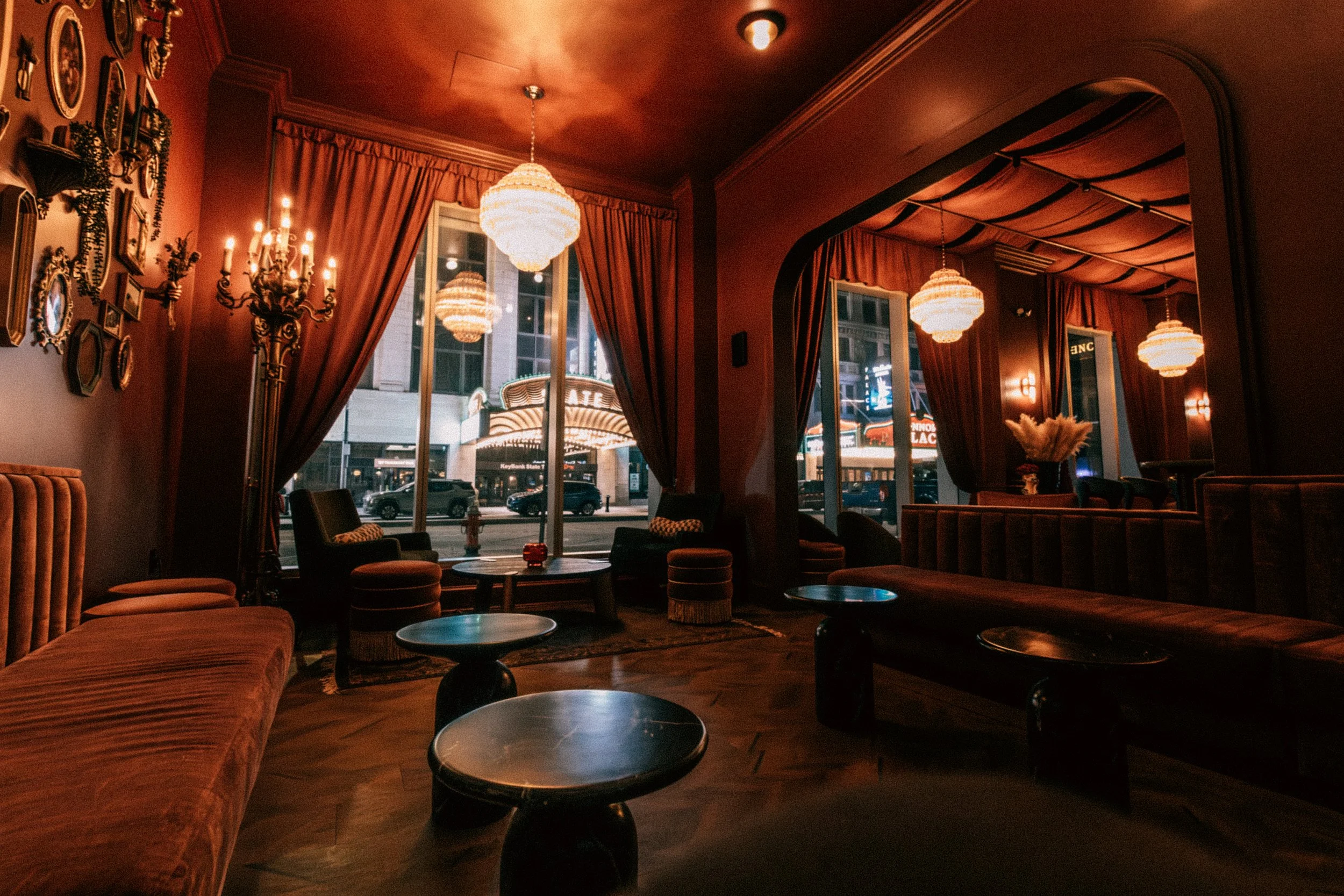 Interior of a vintage-style lounge or bar with dim lighting, velvet seating, round tables, ornate wall décor, large windows with curtains, and city street view at night.