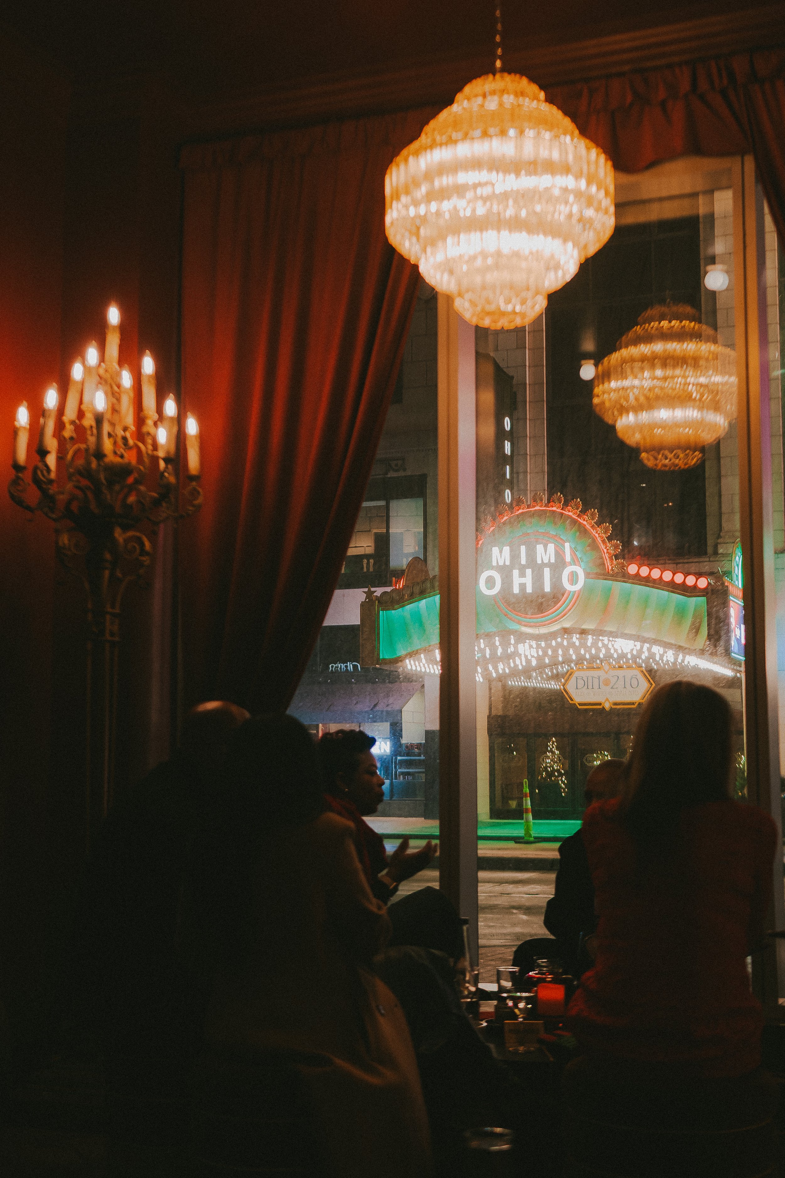 People sitting inside a dimly lit room with elegant chandeliers and red curtains, looking out through glass windows at a neon-lit downtown street scene with theater and signs visible.