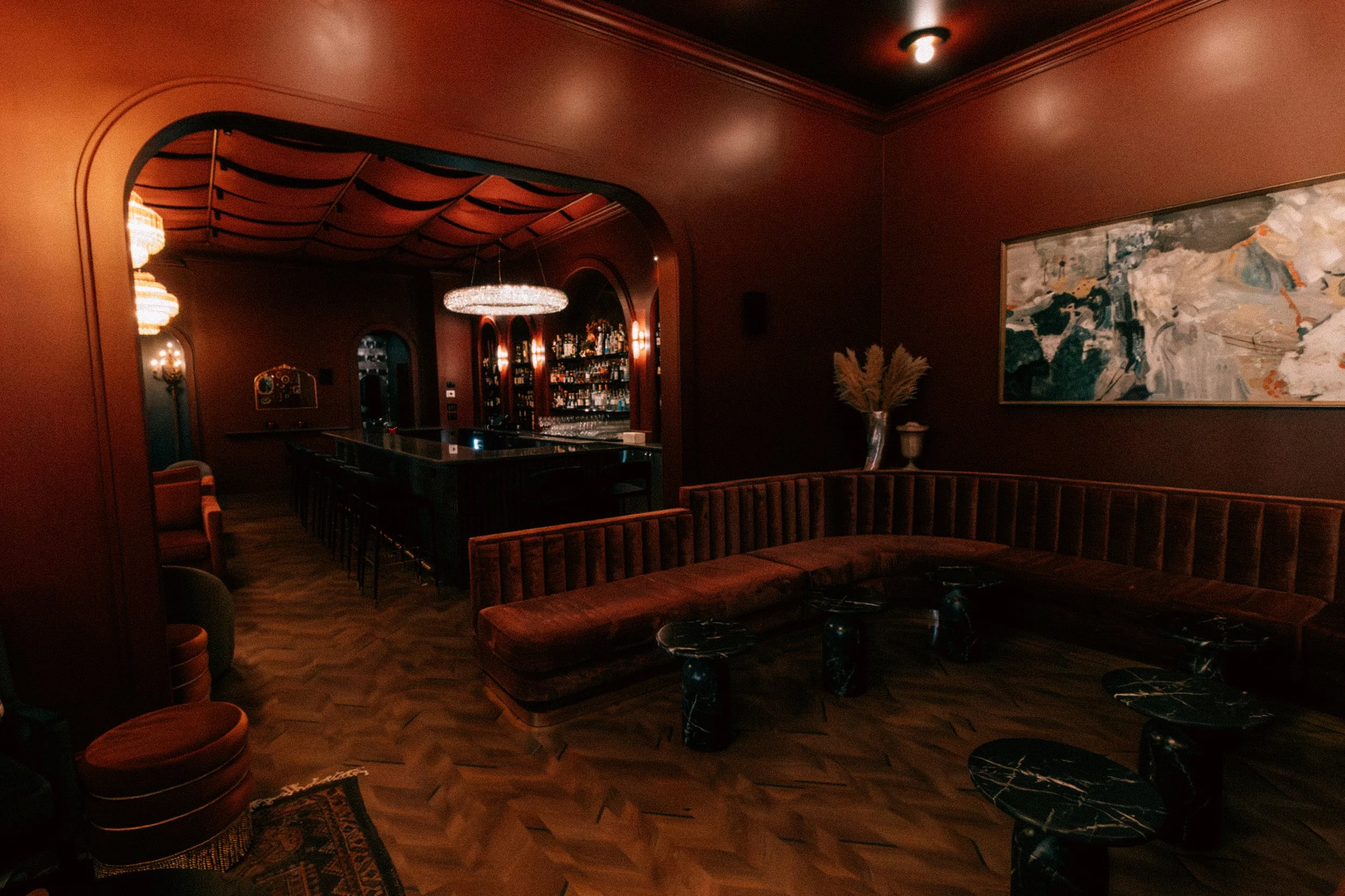 A dimly lit, elegant bar with a curved velvet banquette, small marble-topped tables, and a large abstract painting on the wall. There are warm, ambient lights, and a well-stocked bar in the background.