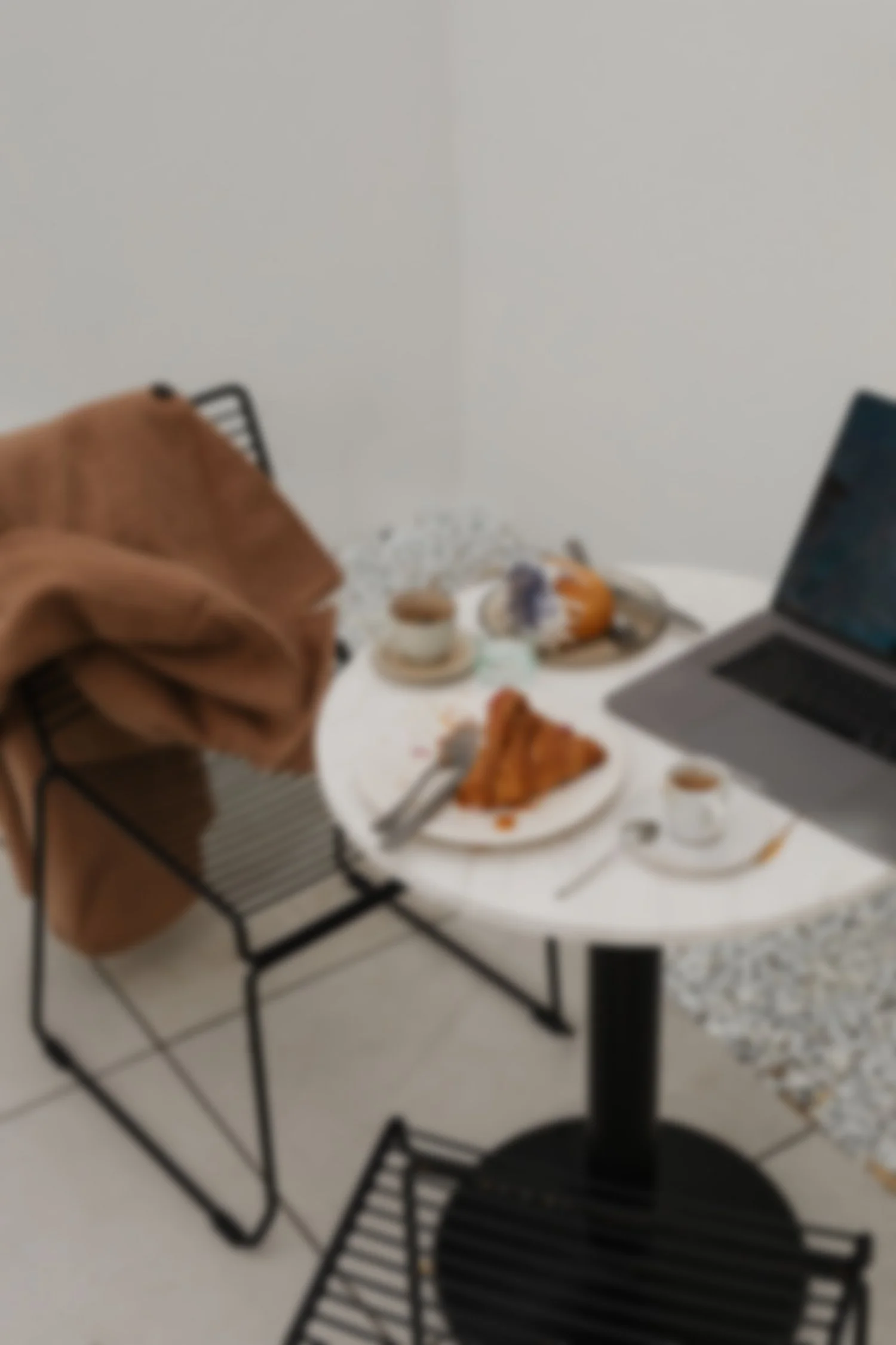 A table with plates of food, including a croissant, and a laptop. There is a chair with a brown coat draped over it and a black dining chair.