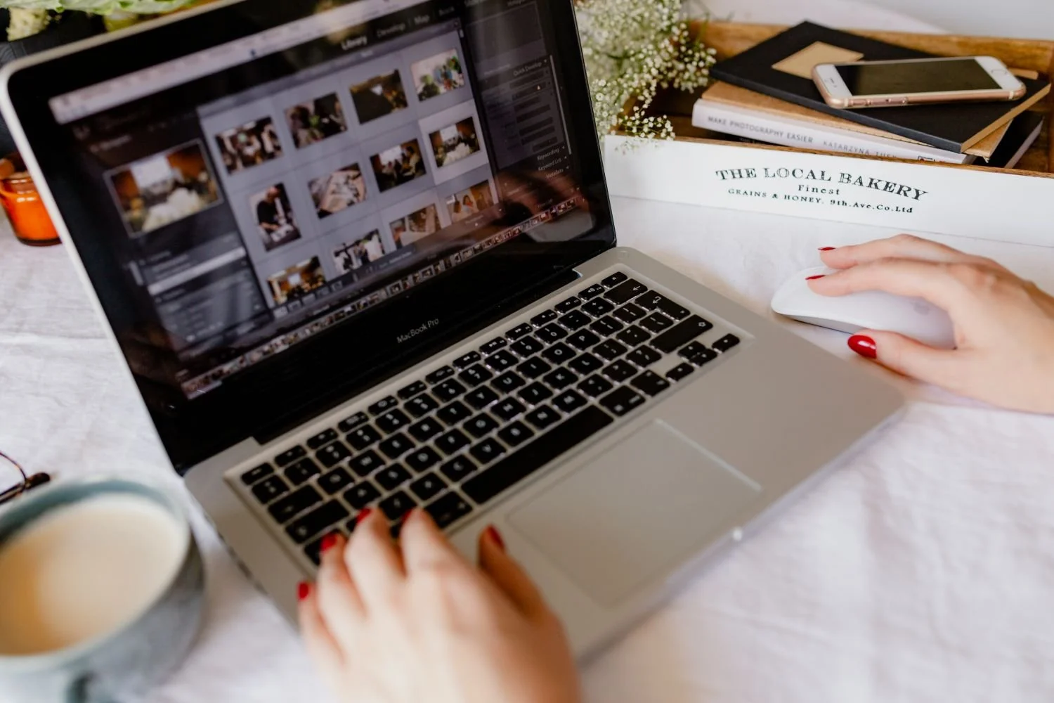A person with red nail polish using a MacBook Pro, editing photos or videos on the screen, with a coffee mug and a smartphone nearby on a desk decorated with flowers and books.