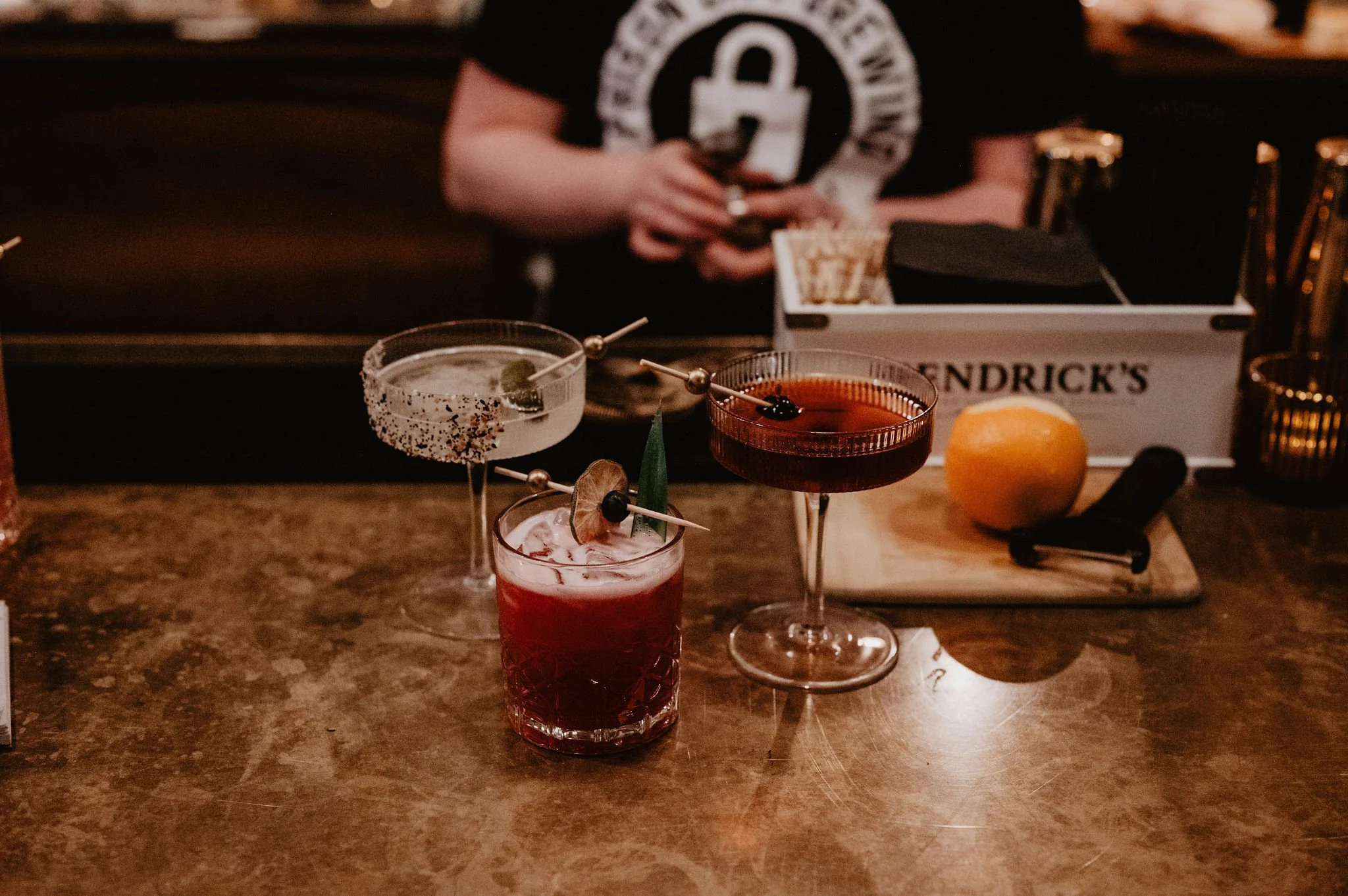 Three cocktails on a bar counter, with a bartender in the background. One with a salted rim and lime, one red colored with dried fruit and a leaf garnish, and one dark red with cherry garnish. There is a lemon and a black bottle opener on a cutting board behind the drinks.