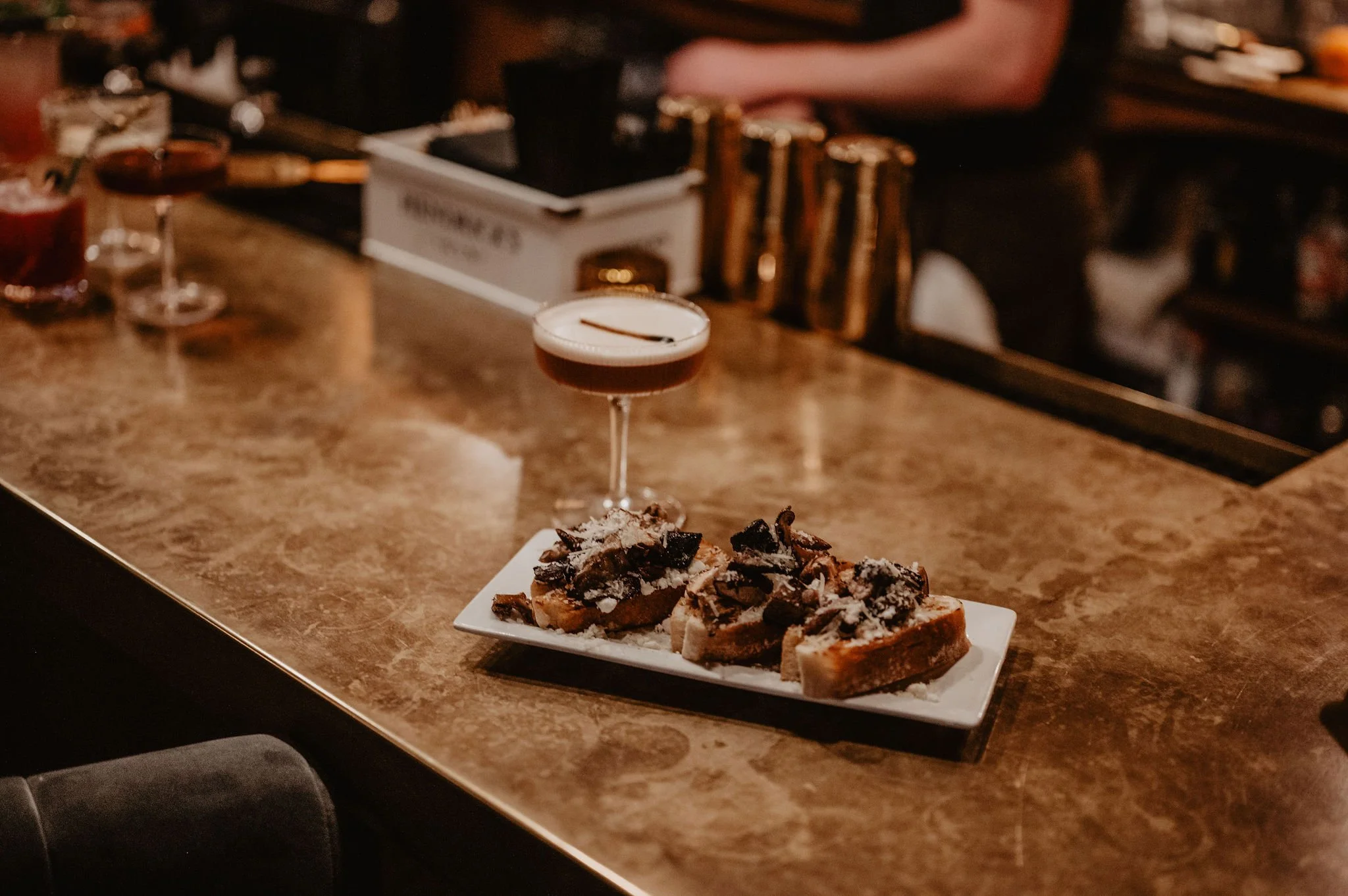 A plate with three pieces of toast topped with chocolate shavings and powdered sugar, alongside a cocktail in a coupe glass on a marble bar counter.