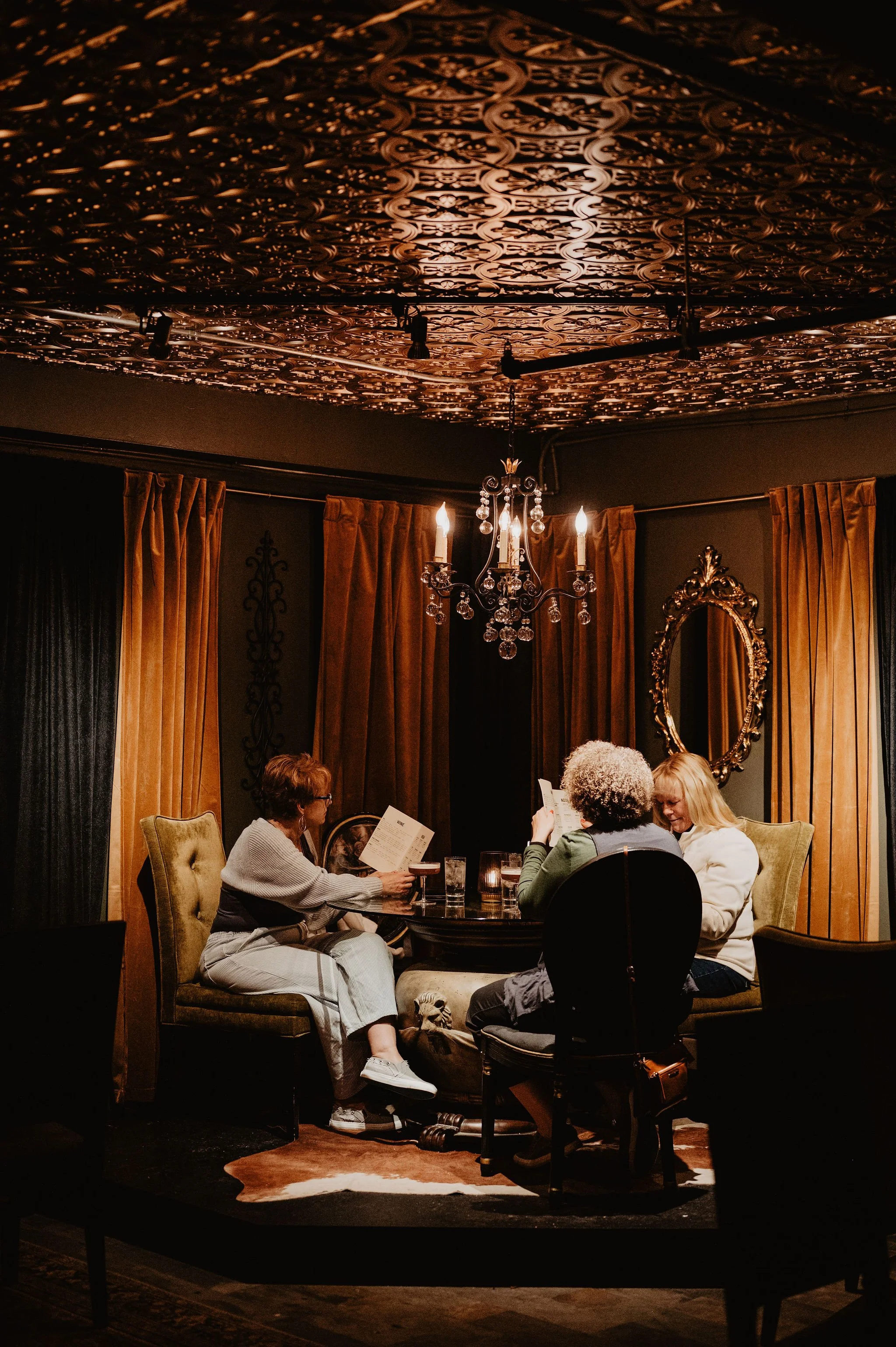 Three women sitting around a small round table in a dimly lit room with ornate decor, reading menus and having drinks.