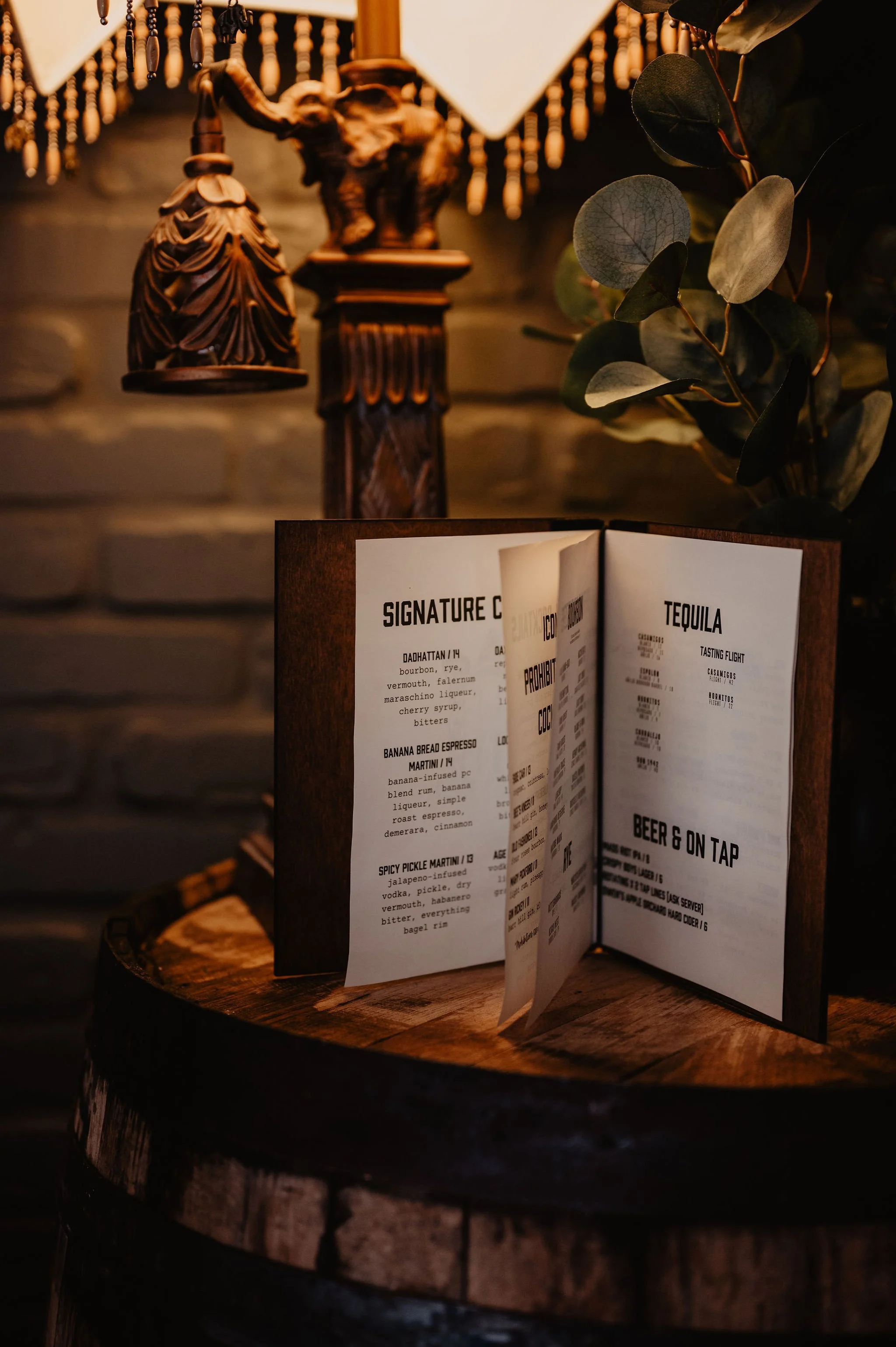 A menu displayed on a wooden stand on a rustic wooden table, with a dimly lit background including a vintage lamp and some greenery.