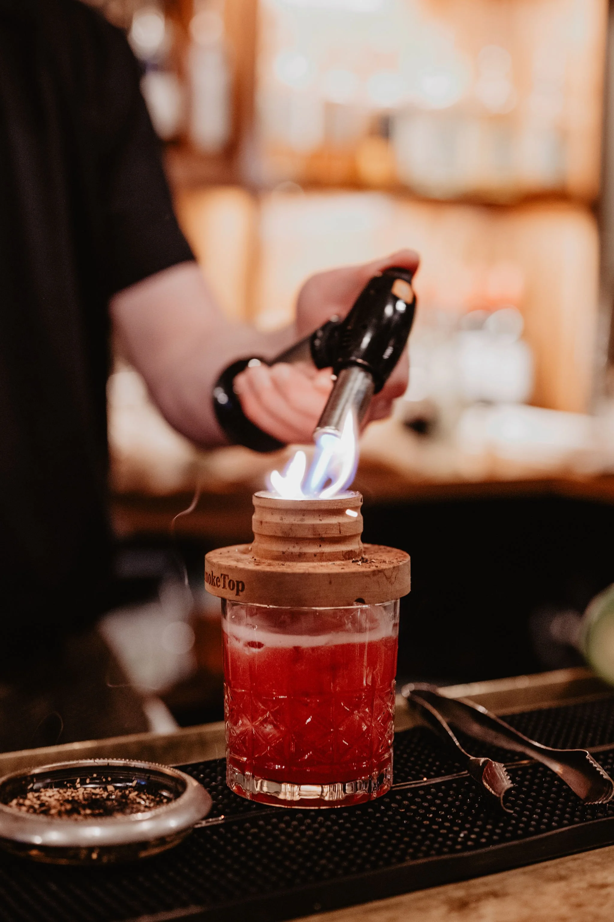 A bartender uses a torch to ignite a flame on a drink in a bar setting.