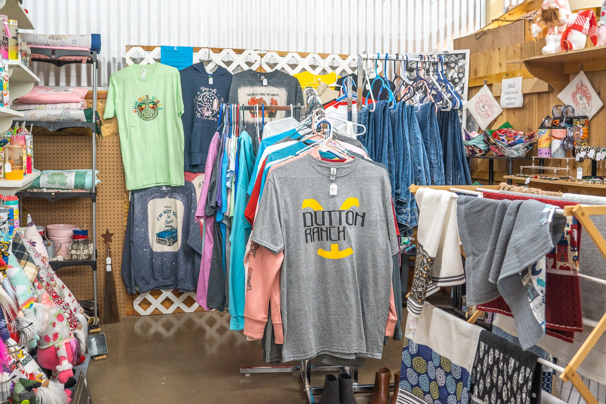 Clothing and accessories displayed on racks and shelves inside a thrift store, including t-shirts, sweatshirts, jeans, towels, and plush toys.