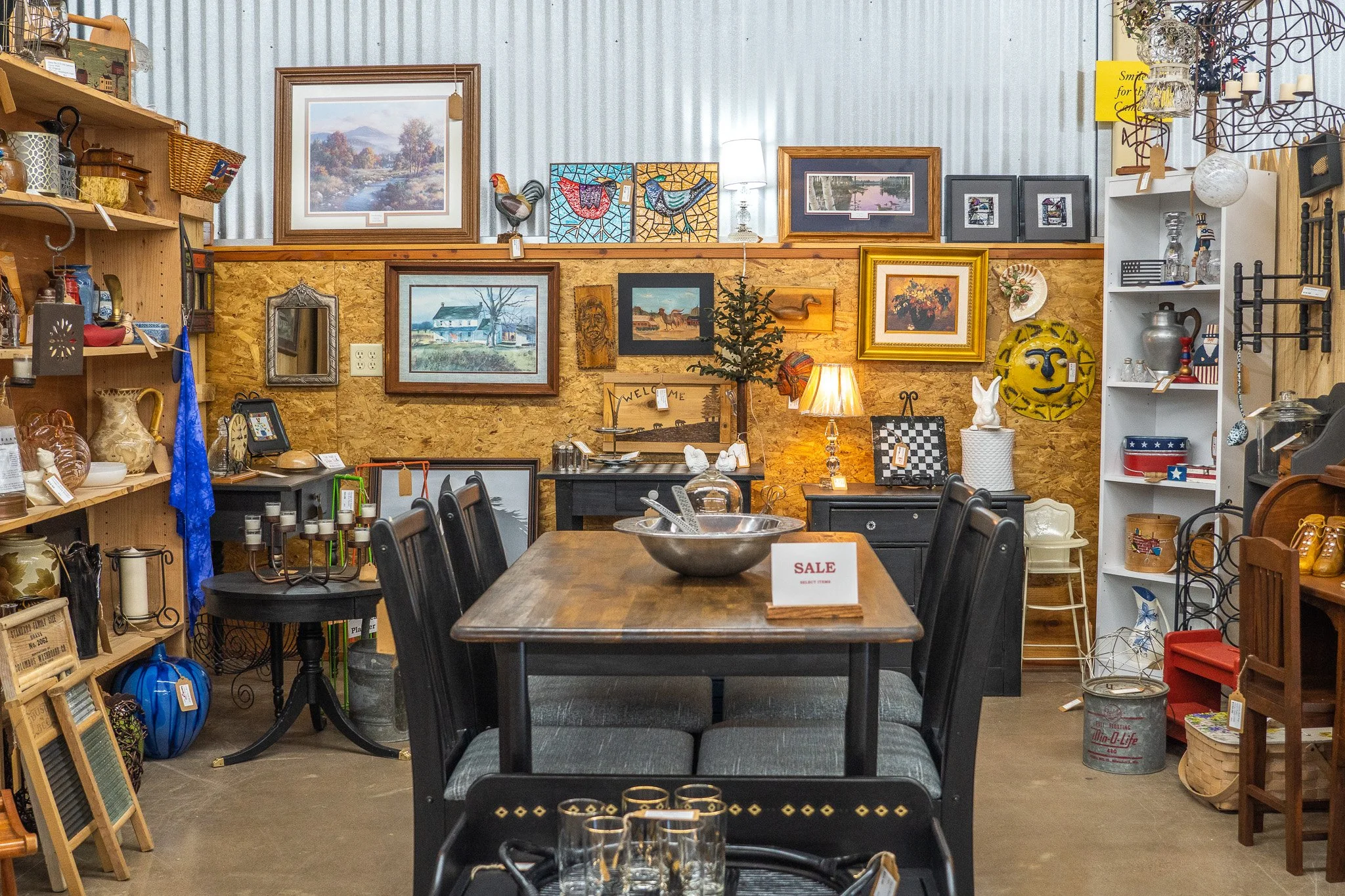 Inside of an antique or thrift store with shelves of decorative items, framed art on the wall, a large wooden table with chairs, and a sale sign on the table.