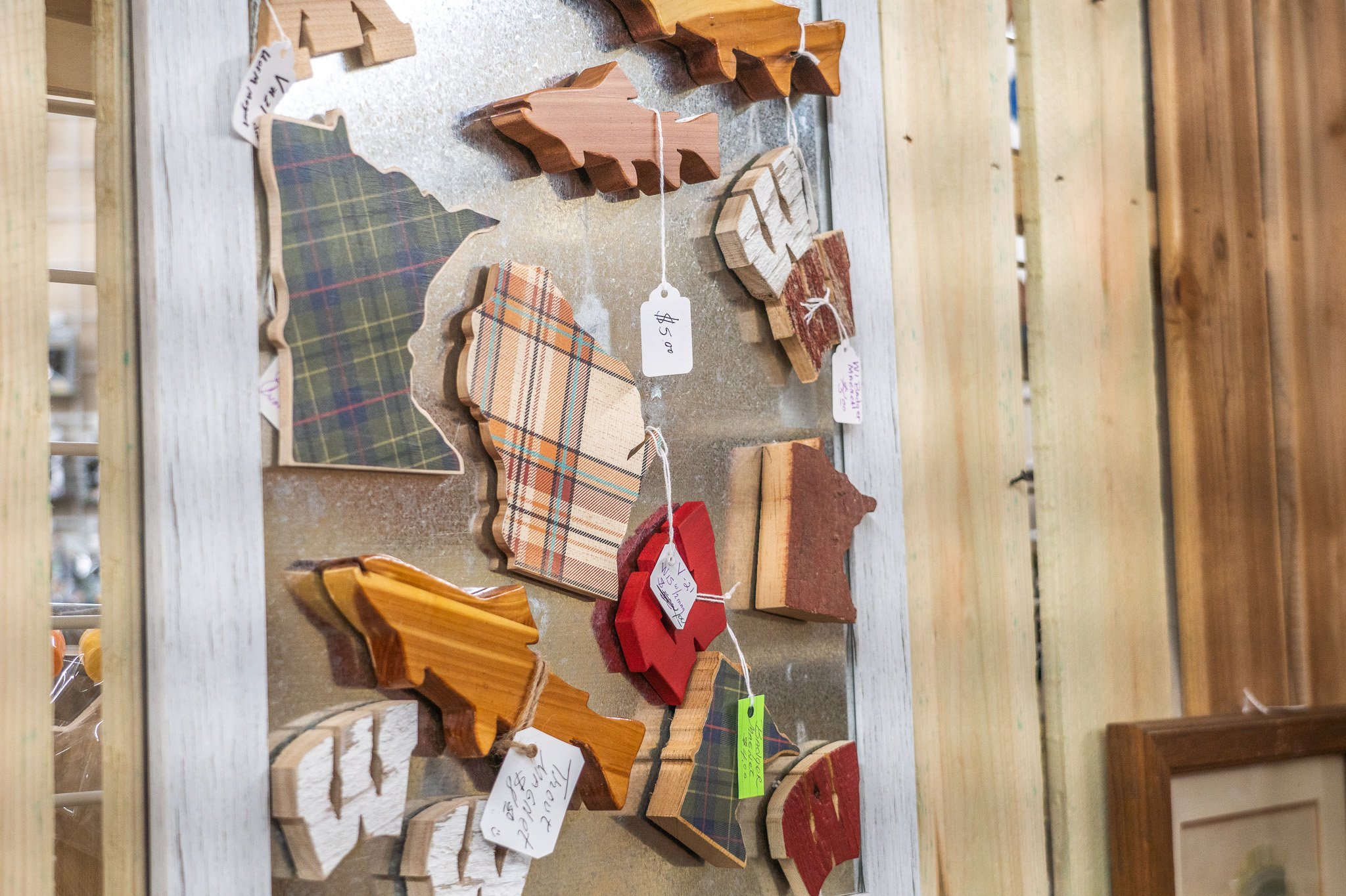 Various wooden Christmas tree ornaments with plaid, white, red, and natural wood finishes displayed on a wooden table, some with price tags.