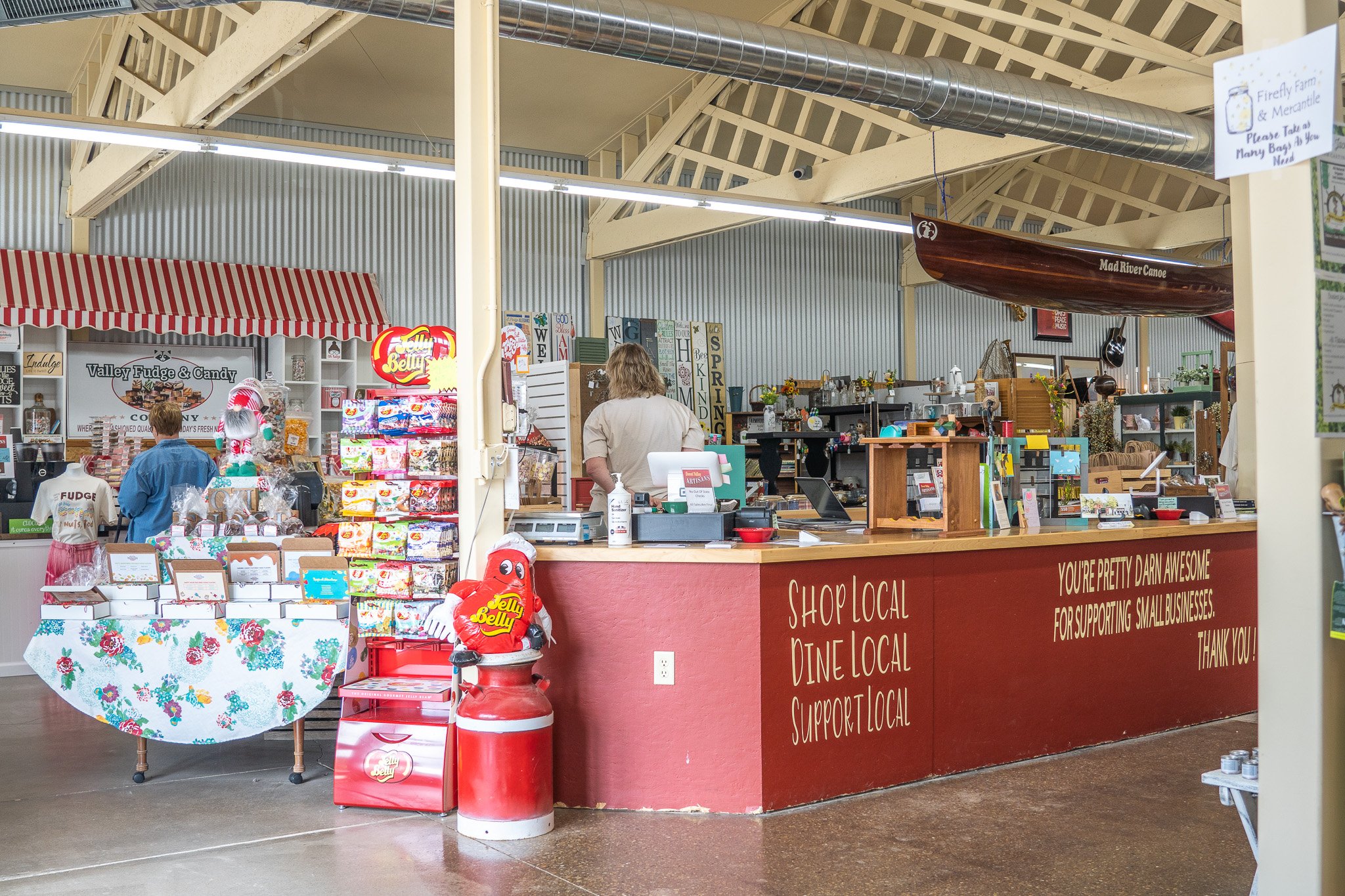 Inside a local shop with a red counter and a sign encouraging supporting small businesses, selling candies, snacks, and crafts, with a woman working behind the counter.