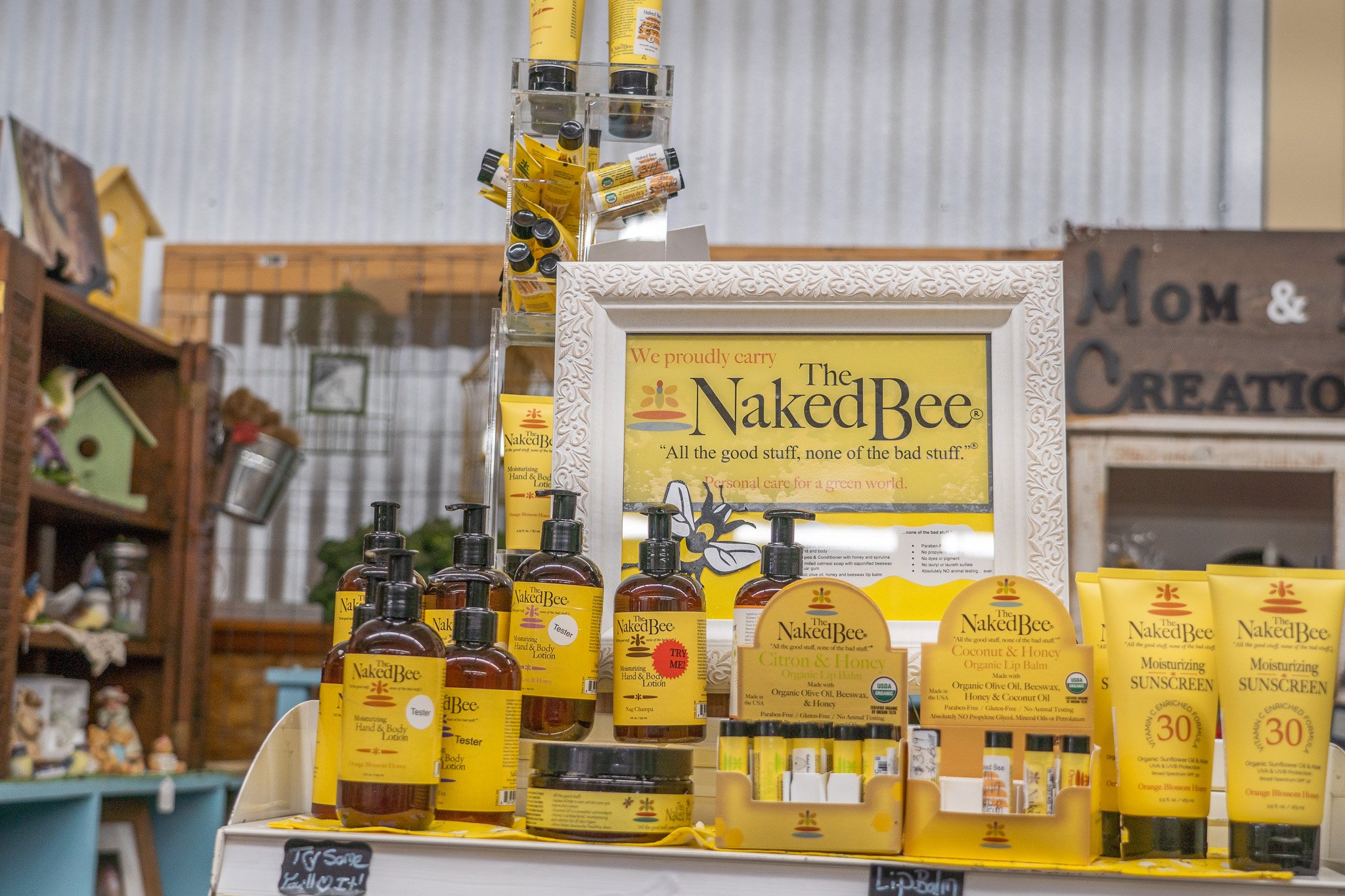 Display of Naked Bee skincare products, including hand and body lotion, lip balm, and sunscreen, on a retail shelf with promotional signage.
