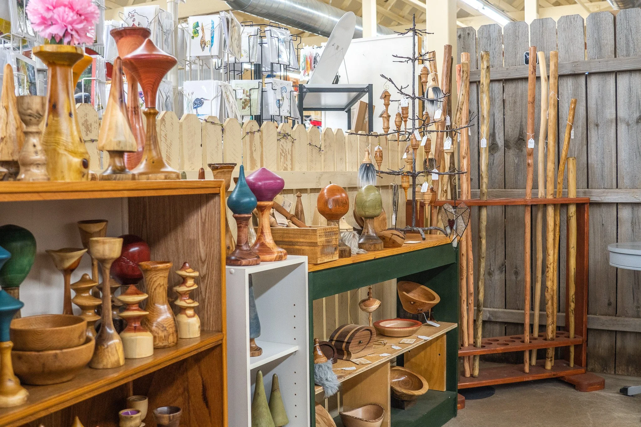 A display of wooden art and home decor items in a store, including vases, bowls, and sculptures made from various woods, arranged on shelves against a wooden fence.