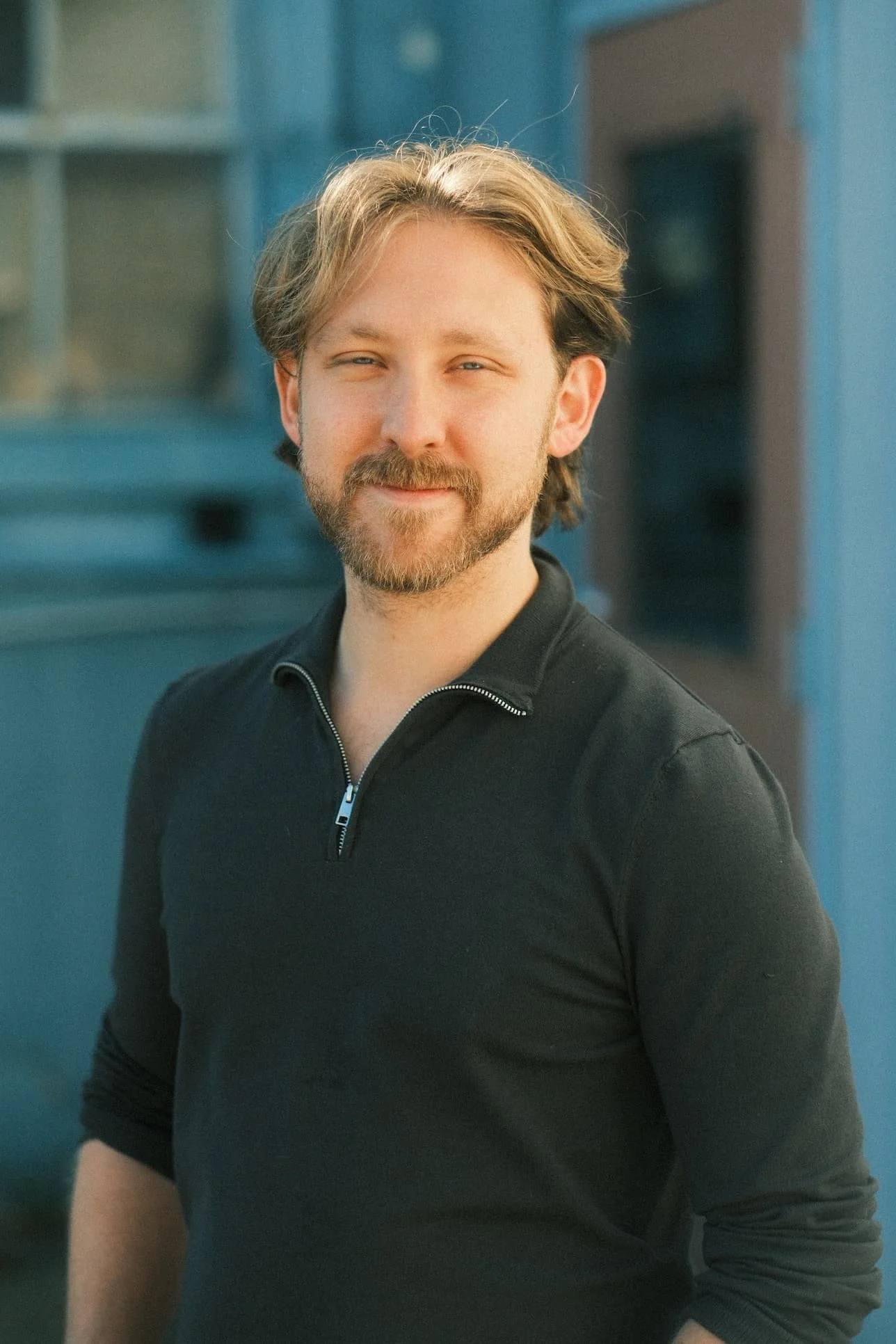 A man with light brown hair and a beard, wearing a black zip-up sweater, standing outdoors in front of a blue building.