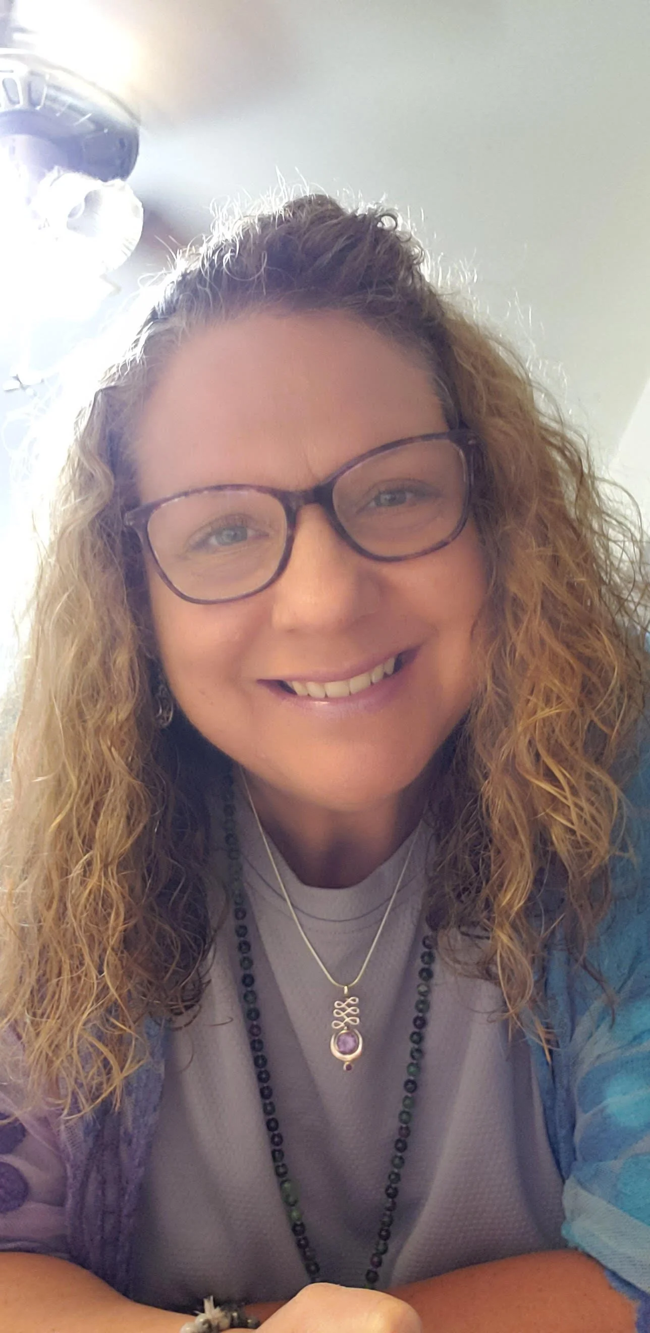 Smiling woman with curly hair, glasses, layered necklaces, and a blue patterned top, sitting indoors.