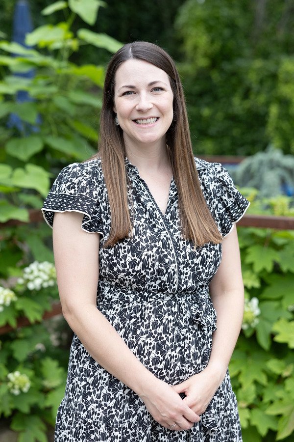 A woman with long brown hair smiling outdoors in front of green leafy plants and flowers, wearing a black and white patterned dress.