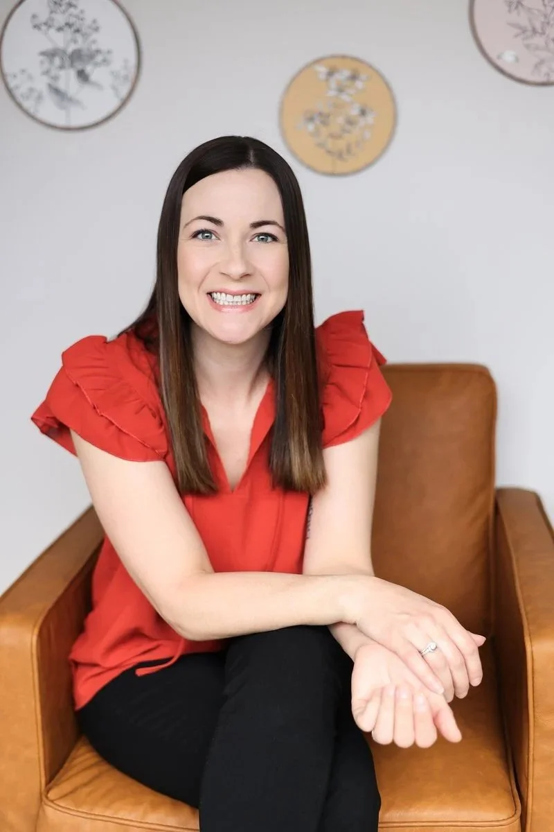 A woman with straight brown hair, wearing a red top with ruffled sleeves, sitting on a tan leather chair, smiling, with decorative wall art behind her.
