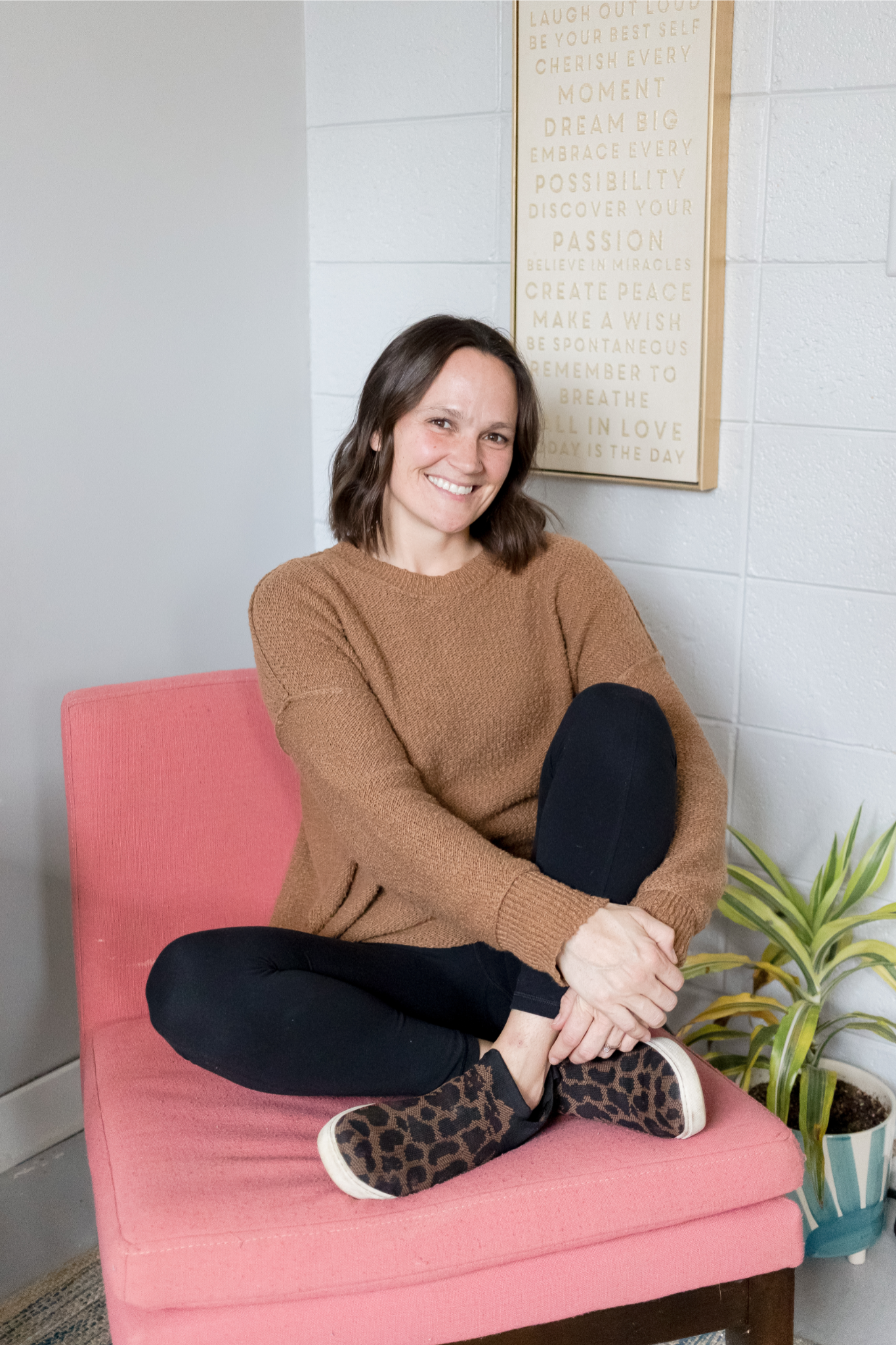 A woman sitting cross-legged on a pink upholstered chair, smiling, wearing a brown sweater, black pants, and leopard-print shoes, with a potted plant and a motivational quote wall art in the background.