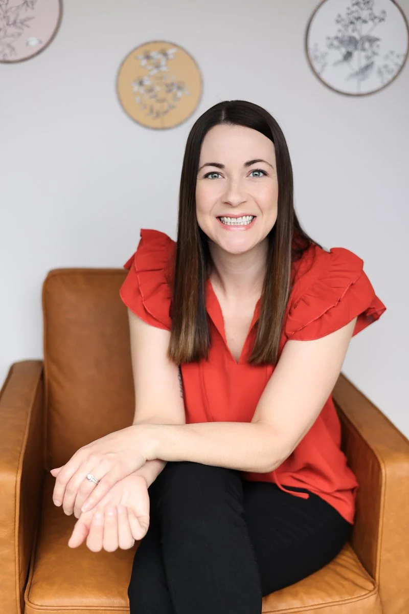 A woman with long brown hair smiling outdoors in front of green leafy plants and flowers, wearing a black and white patterned dress.