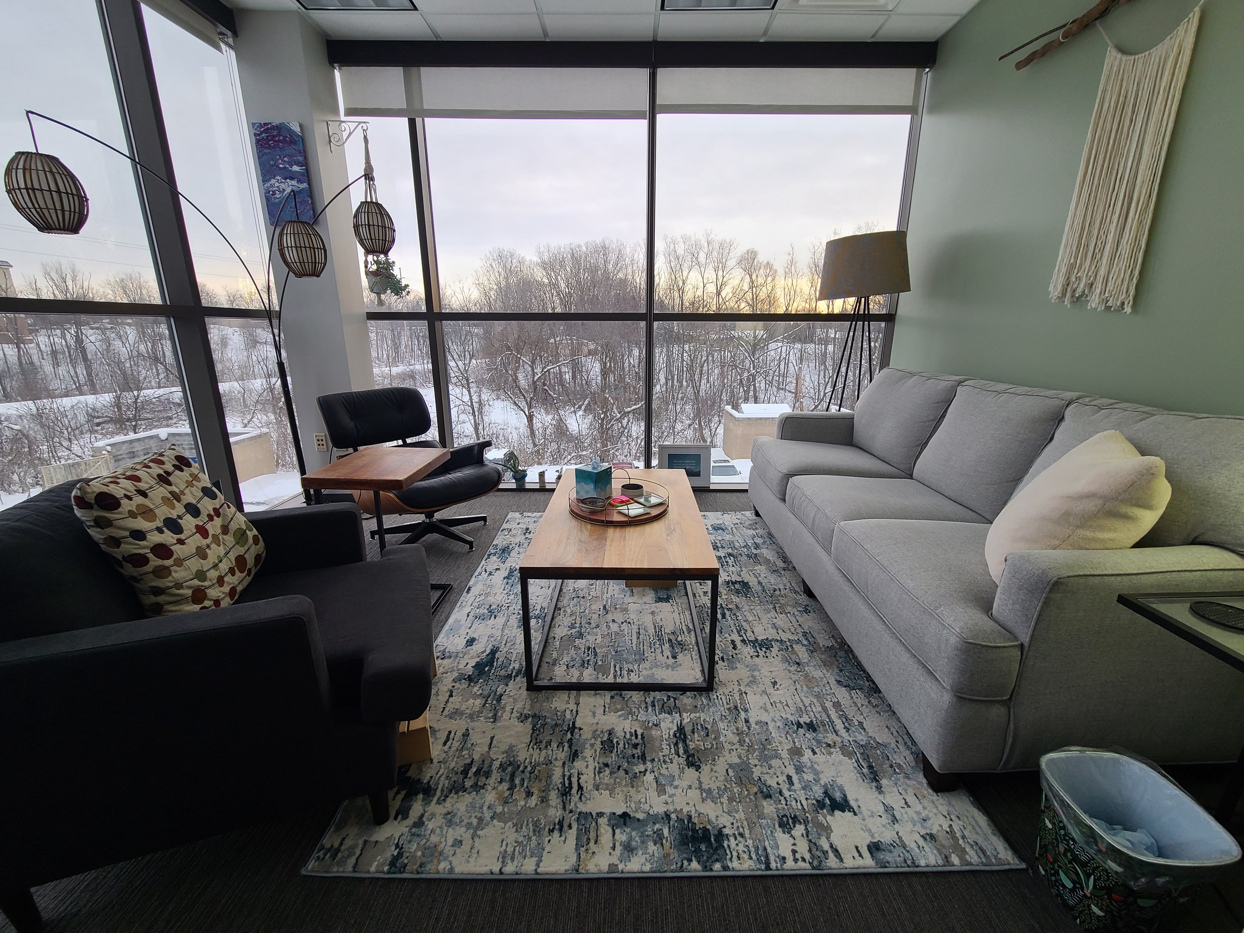 Living room with large windows overlooking snowy trees, furnished with a gray sofa, black armchair, wooden coffee table, and decorative hanging lamps.