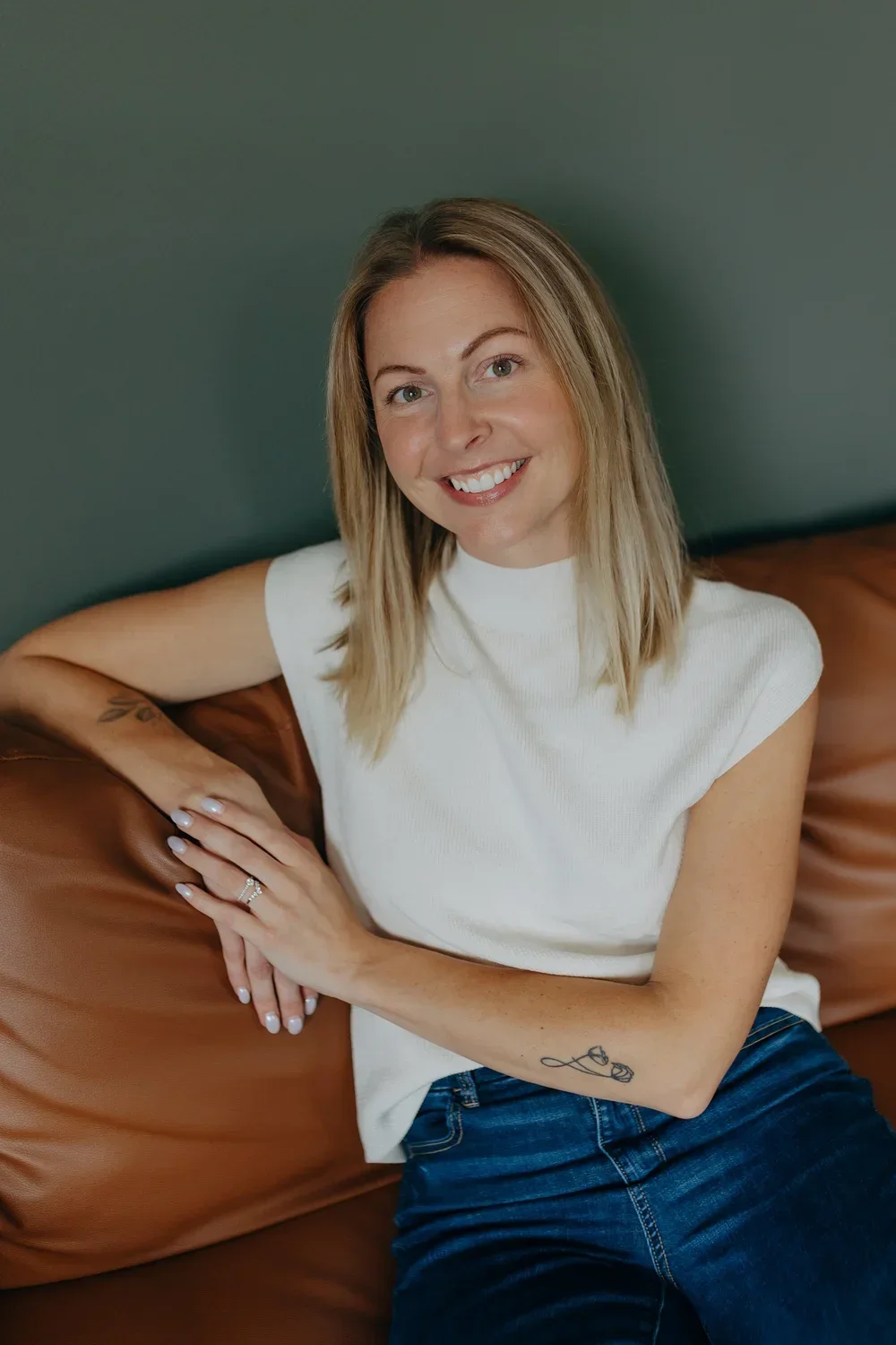 A woman with shoulder-length blonde hair, smiling, sitting on a brown leather couch, wearing a white t-shirt and blue jeans, with tattoos on her left arm and right wrist, against a dark green wall.