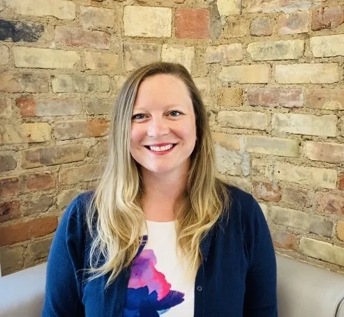 A woman with blonde hair smiling, wearing a white shirt with a colorful abstract design and a blue cardigan, sitting against a brick wall.