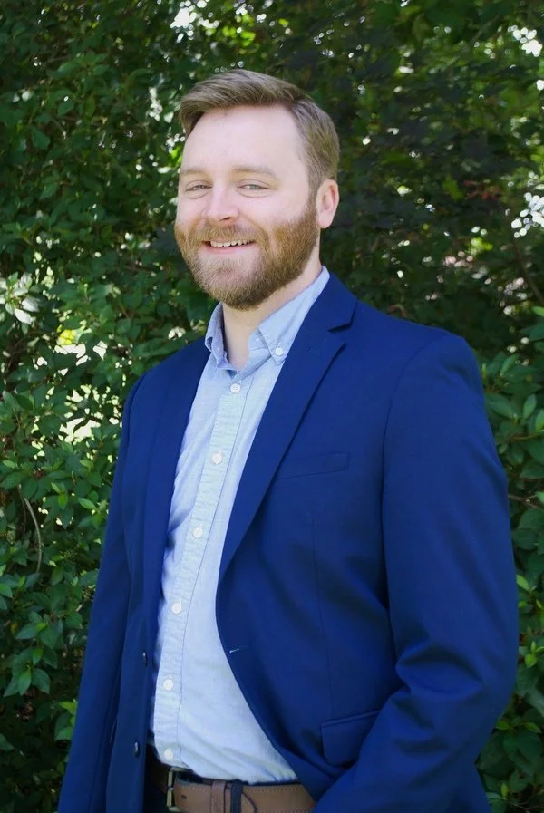 A young man with light brown hair and a beard, wearing a blue blazer and a light blue shirt, smiling outdoors with green foliage in the background.