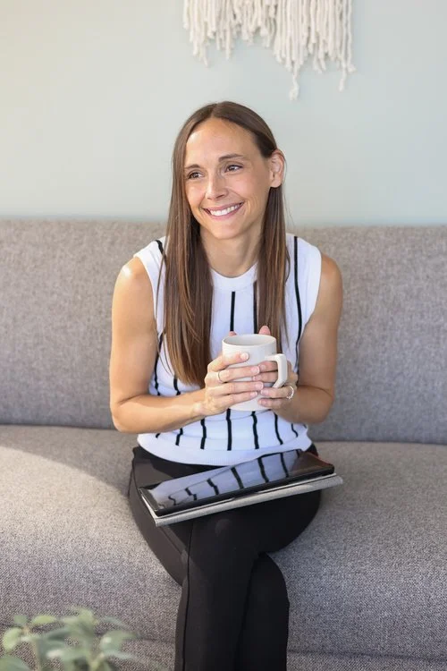 A woman sitting on a gray couch, holding a mug and smiling, with a tablet resting on her lap.
