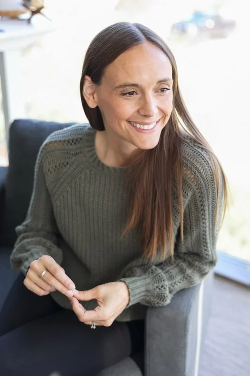 Tracy Frostick, A woman smiling while sitting on a couch in a well-lit room with a large window behind her.
