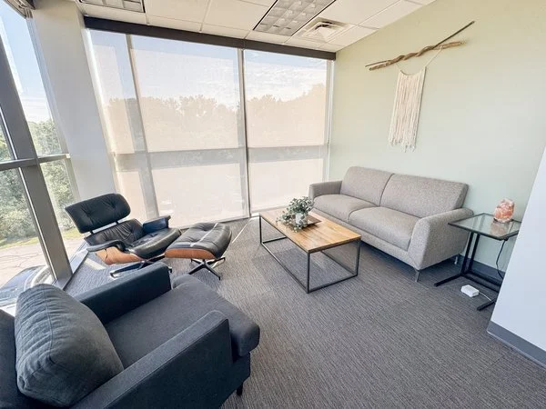 Office lounge with a gray sofa, black armchair, Eames lounge chair, coffee table, side table, and large window with blinds.