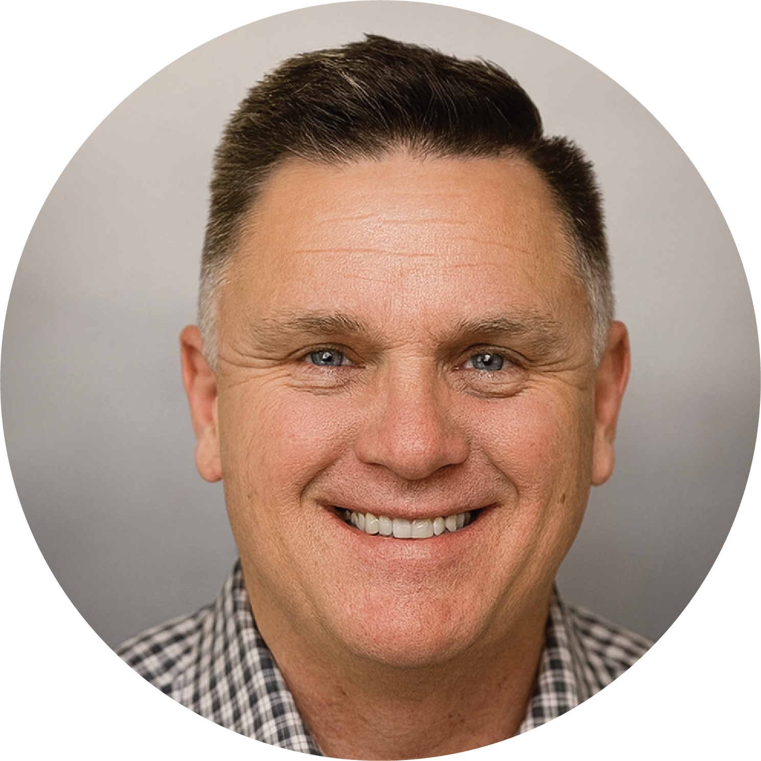 A professional headshot of a man with short brown hair, blue eyes, wearing a checked shirt and blazer, smiling against a gray background.