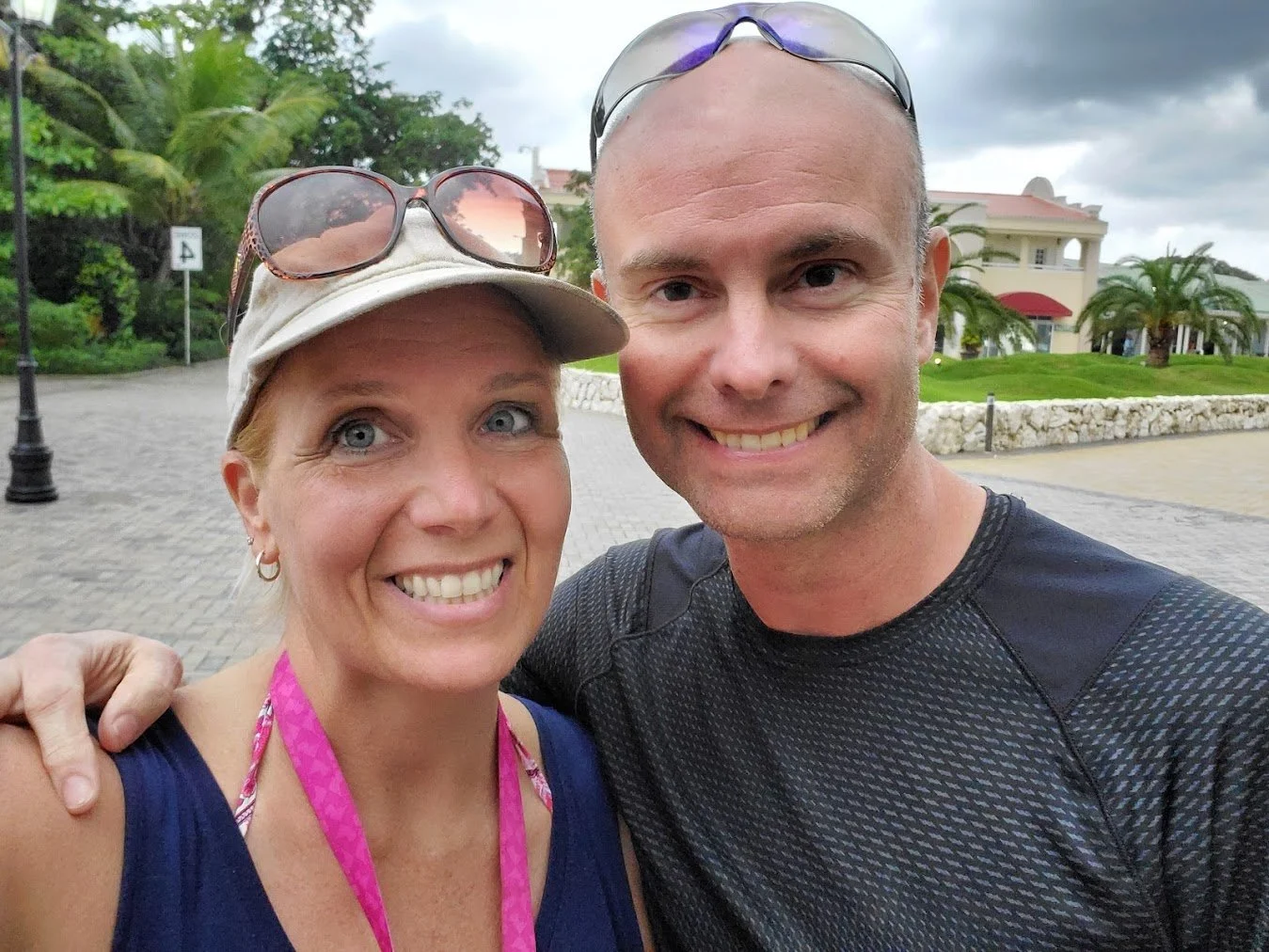 A smiling man and woman taking a selfie outdoors in front of a building with palm trees. The woman is wearing a beige cap, sunglasses on her head, and a blue tank top. The man is wearing sunglasses on his head and a black athletic shirt.