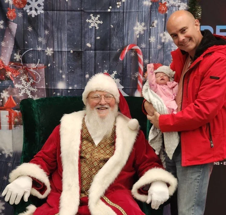 A man dressed as Santa Claus sitting on a green chair, smiling, with a man holding a baby girl dressed in pink next to him, in front of a festive Christmas-themed backdrop.