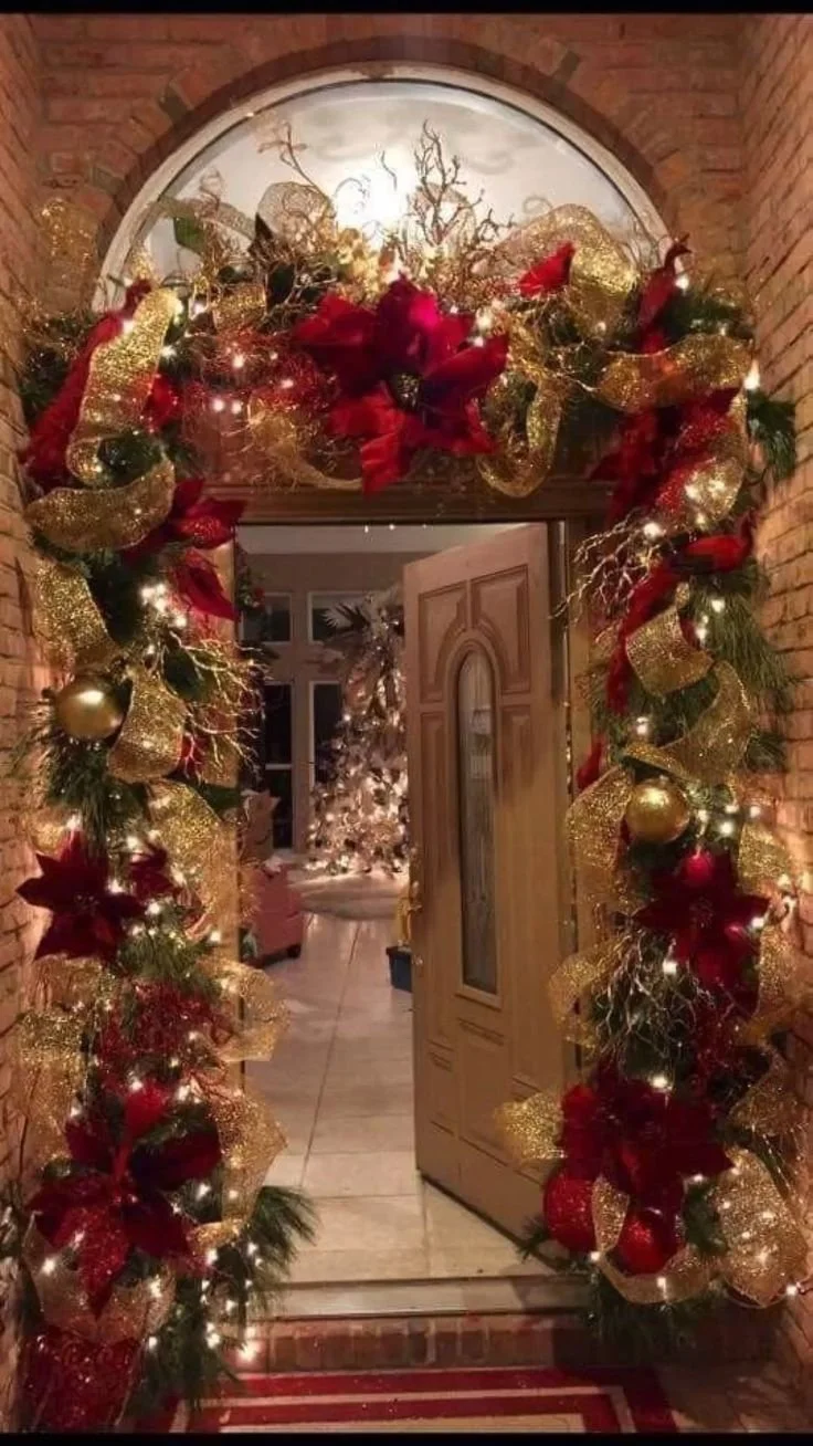 Decorated Christmas archway with gold and red ornaments, ribbons, and lights, leading into a home with a Christmas tree visible inside.