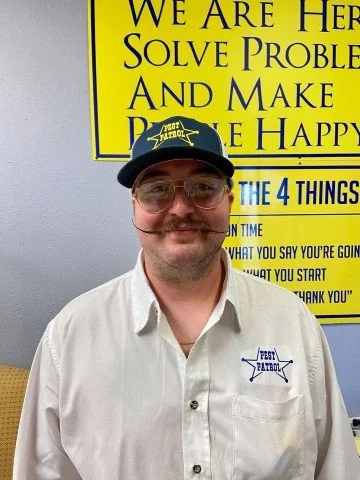 man in a pest patrol shirt and hat standing in front of a yellow sign with blue writing. 