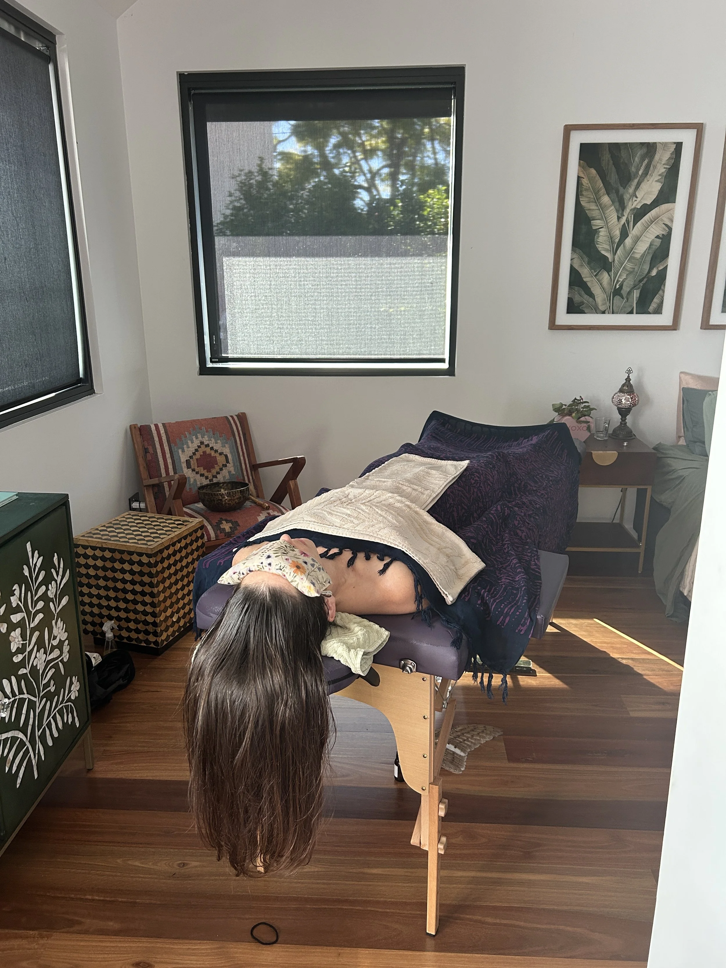 A woman with long brown hair lying face-down on a massage table in a room with wooden floors, framed pictures of leaves on the wall, and a window showing trees outside.