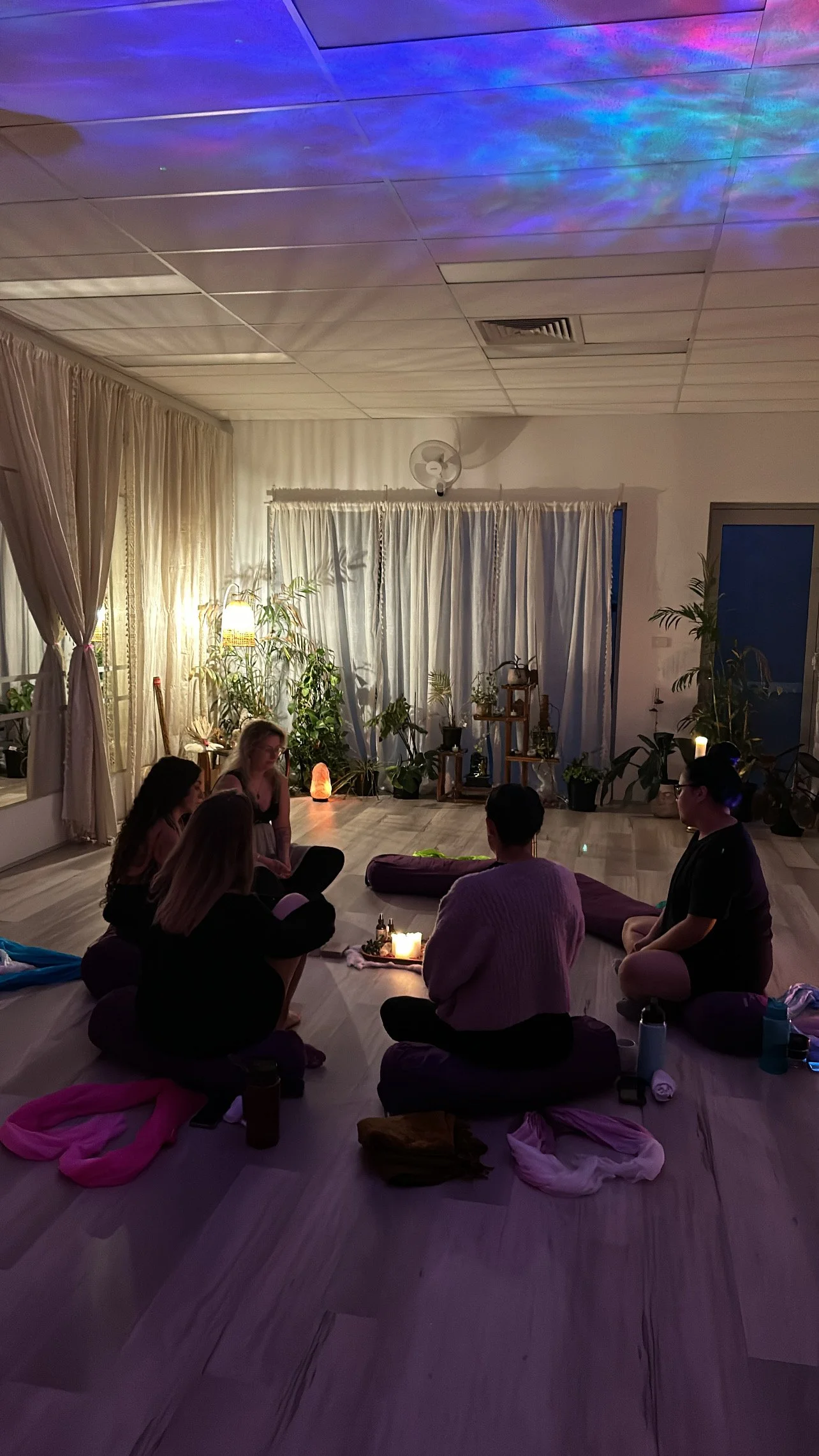 A group of people sitting cross-legged around a candle-lit circle in a dimly lit room decorated with plants, curtains, and soft ambient lighting, possibly engaged in a meditation or yoga session.