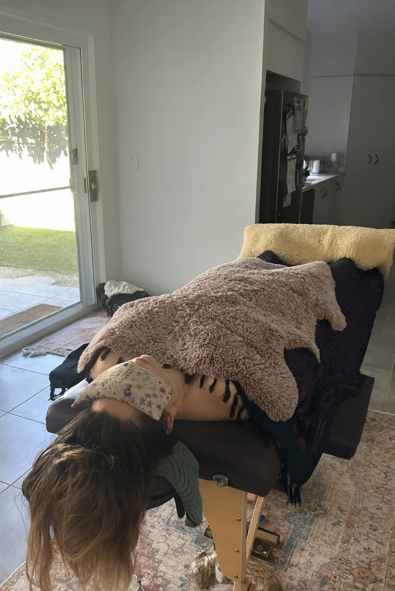 A person is lying on a massage table with a blanket covering them, a woman with long hair is resting on the table, a cloth with a floral pattern is covering her face, and a small dog stands underneath the table near her feet. The room has a sliding glass door with sunlight coming in, and a kitchen can be seen in the background.