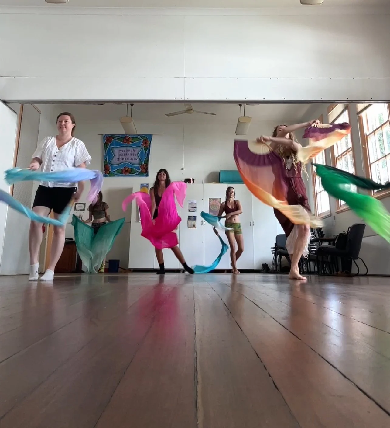 Group of women practicing silk fans in a bright studio with wooden floors and large windows.