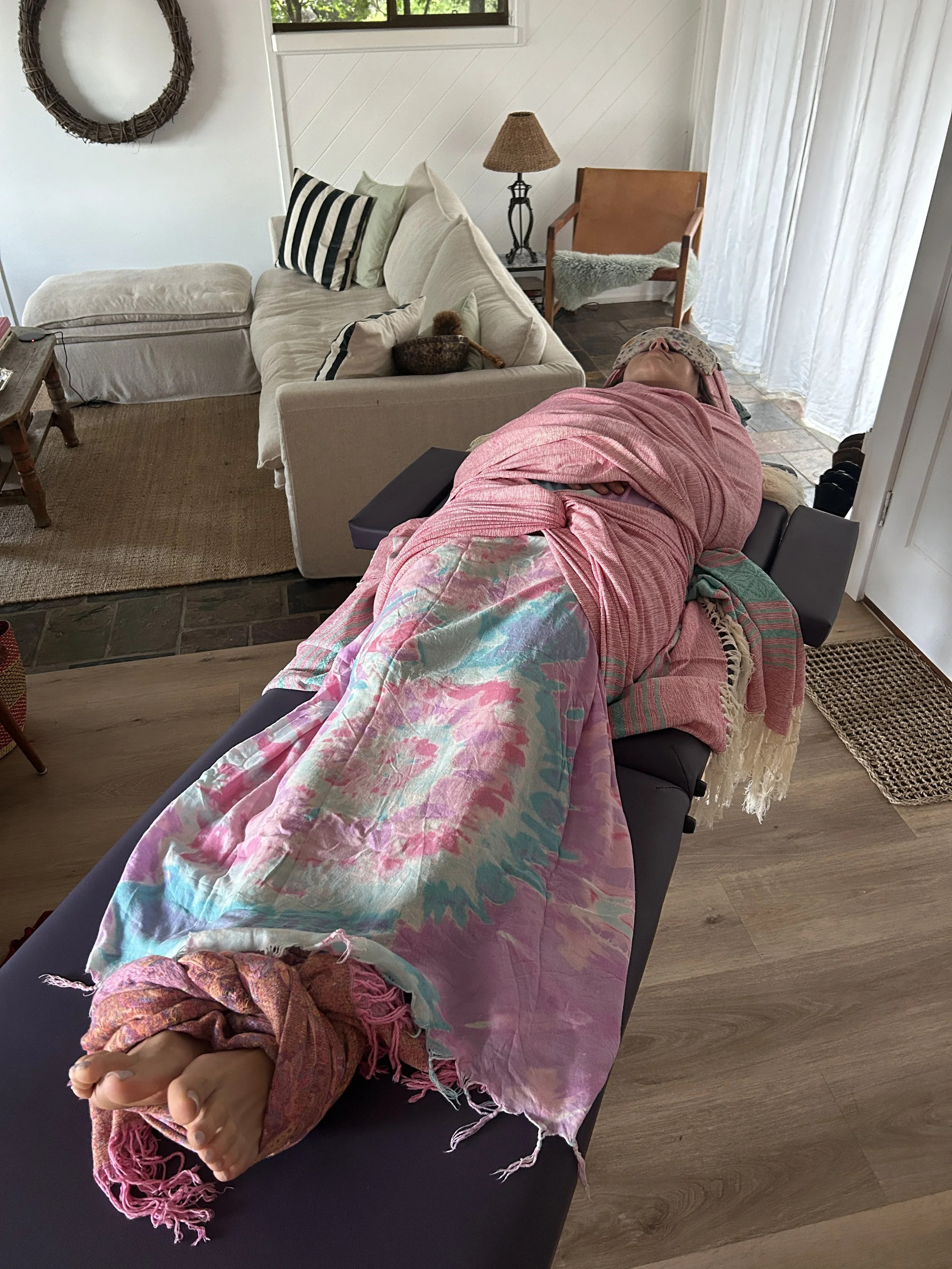Person resting on a therapy table covered with Rebozo, with feet visible at the foot of the table. The room has a beige sofa with pillows, a side table, a lamp, a wooden chair, and a window with white curtains.
