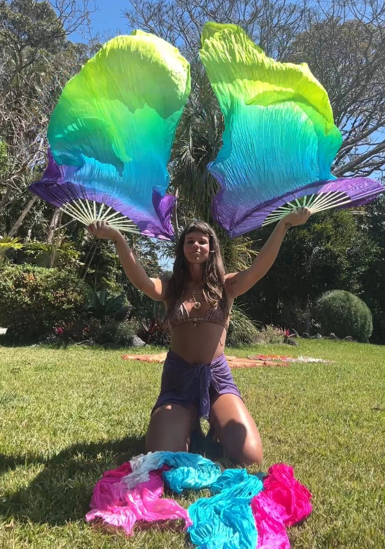 A woman with long brown hair kneels on grass outdoors, holding large rainbow-colored silk fans open above her head. Colorful fabric pieces are on the ground around her, and trees and bushes are visible in the background under a clear blue sky.