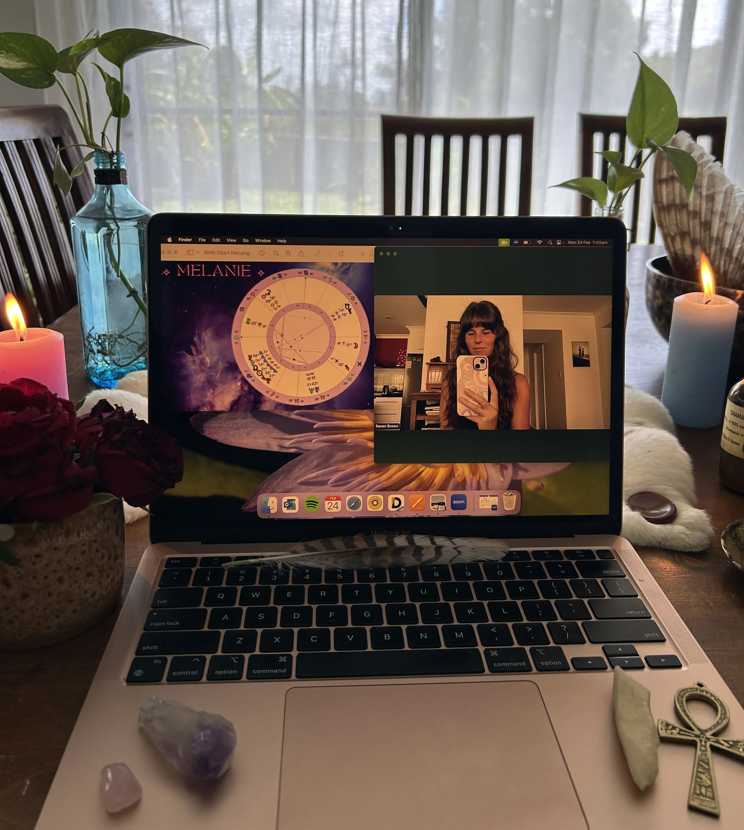 A laptop on a wooden dining table displays a birth chart for Melanie and a video call window showing a woman taking a selfie. The table is decorated with candles, flowers in vases, rocks, and an ankh cross.