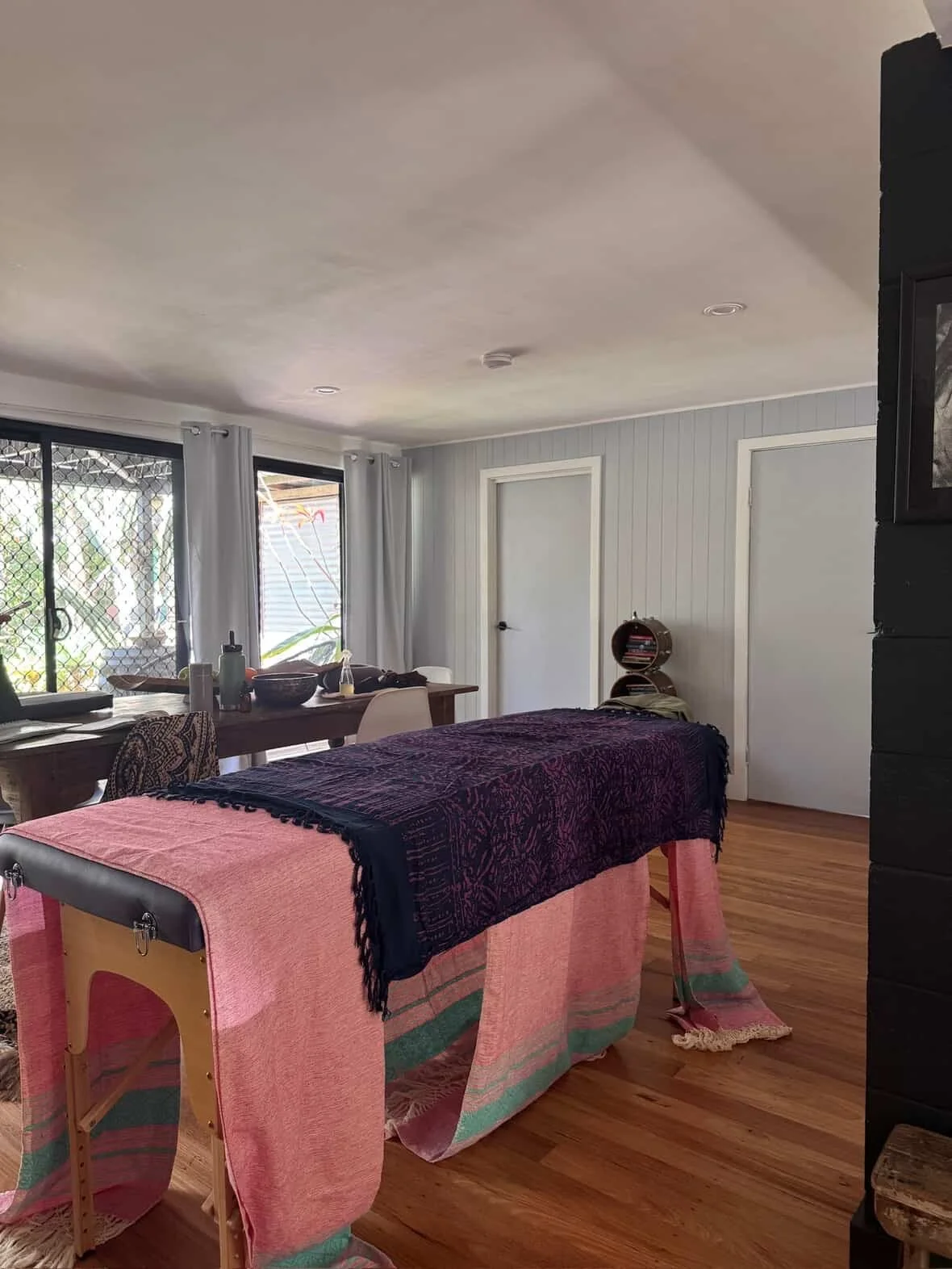 A room with a massage table covered with cloths, a dining table with chairs nearby, windows with white curtains, and wooden flooring.