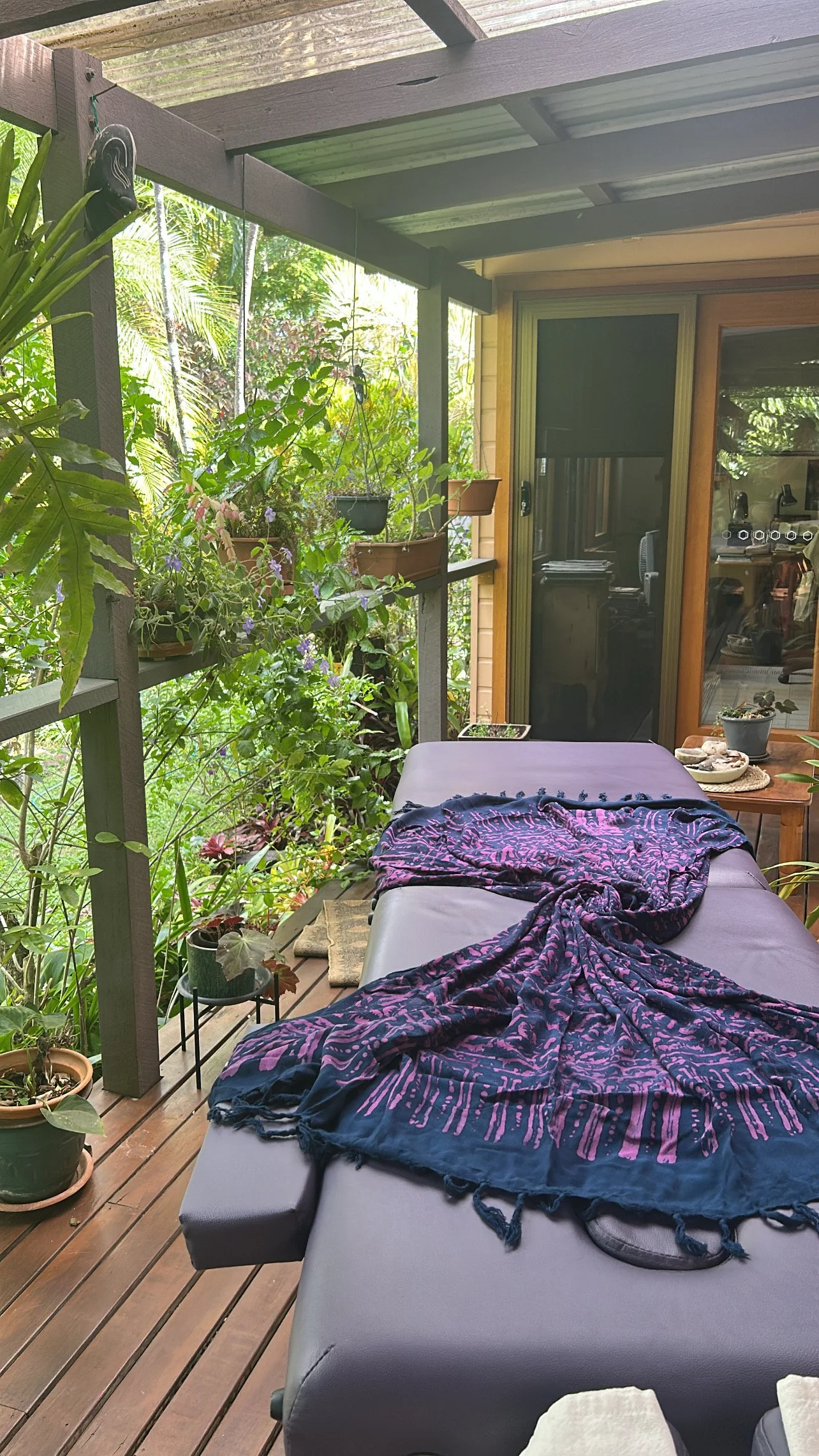 A balcony with wooden flooring, decorated with plants hanging and sitting in pots. There is a massage table with a purple and pink patterned cloth draped over it, and a small side table with a pot of plants and some food items. Doors lead into the house with a visible interior.