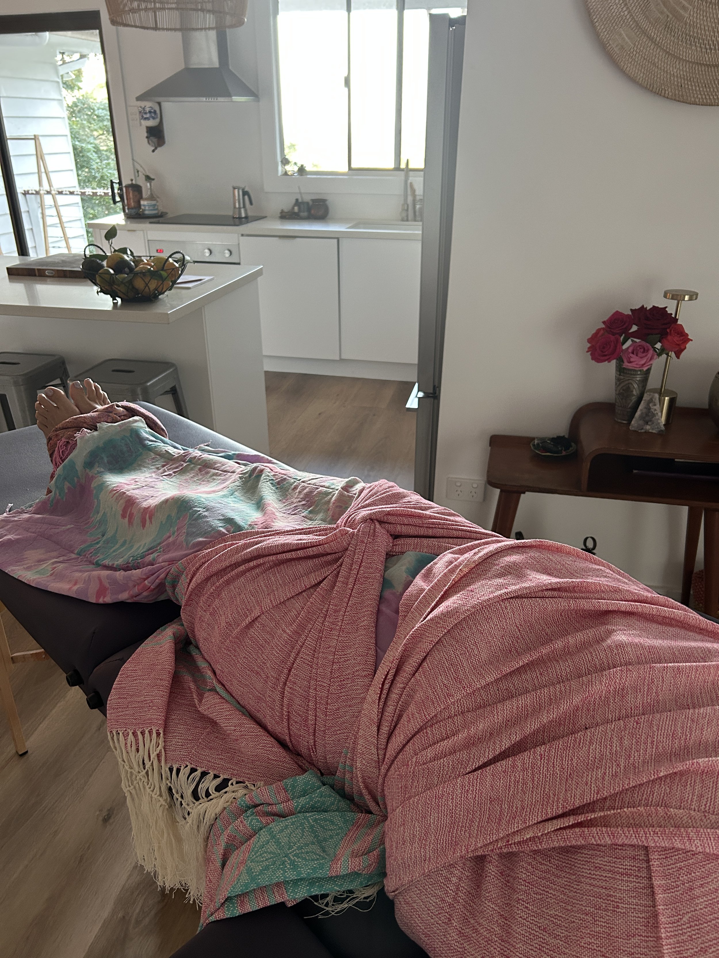 Person lying on a massage table covered with a pink, blue, and green blanket, in a room with a wooden floor and a small table with a vase of red and pink roses, next to a wall with a woven wall hanging. The background shows a kitchen area with a white island, a fruit bowl, and a window.