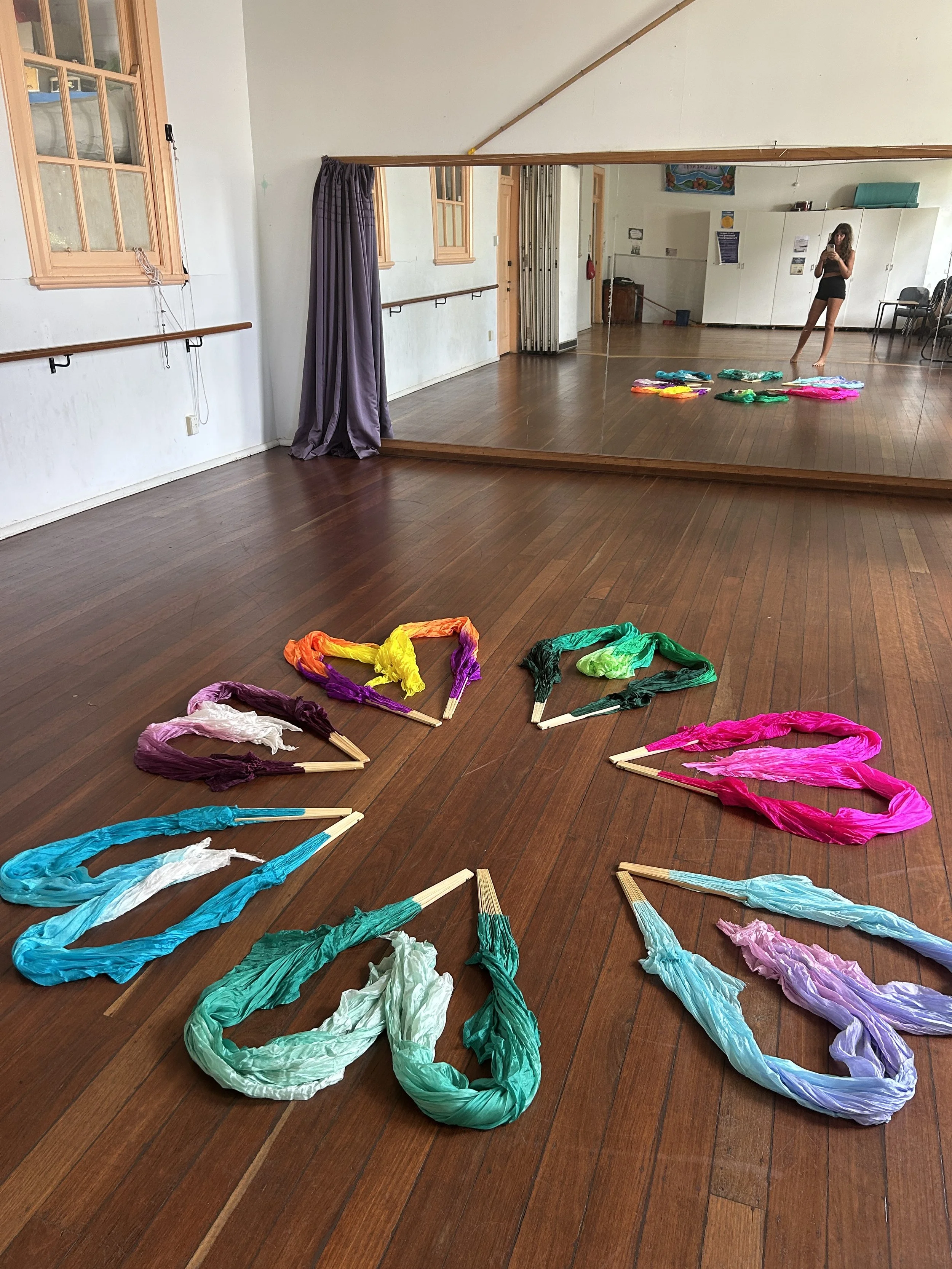 Colorful silk fans arranged in a semi-circle on a wooden dance studio floor with a large mirror reflecting the room and a person taking a photo.