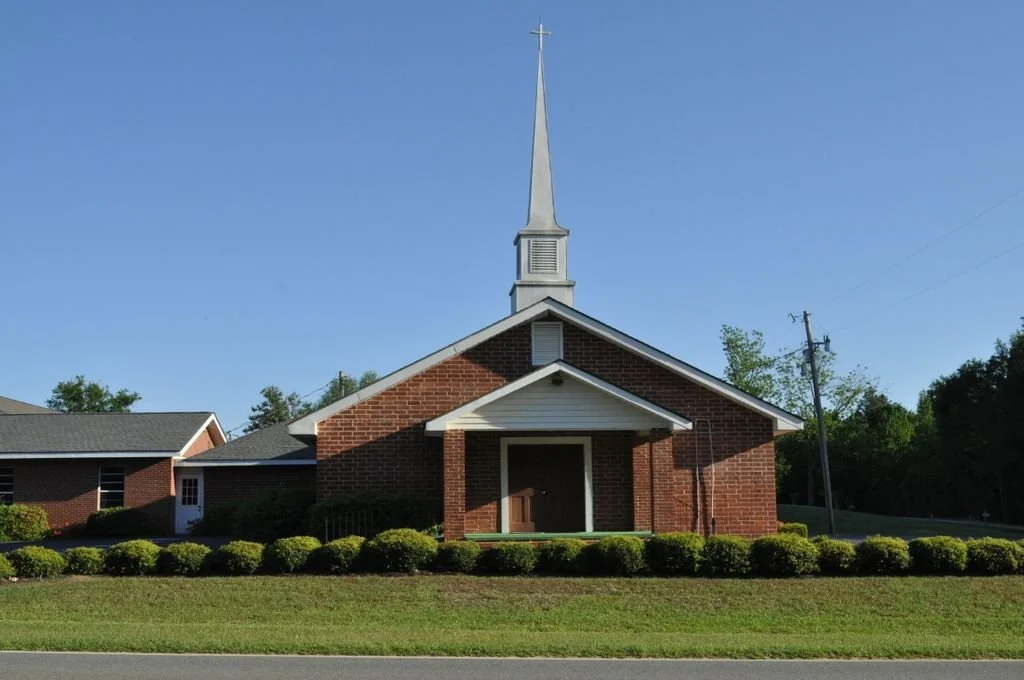 A small brick church with a white steeple topped with a cross, surrounded by green bushes and trees, under a clear blue sky.