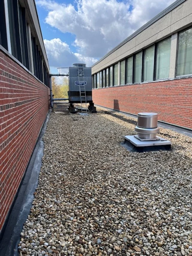 Gravel flat rooftop with HVAC unit, and vent, between two brick buildings under a partly cloudy sky on an industrial building in New England. WP Walsh Roofing got and installed all the materials needed.